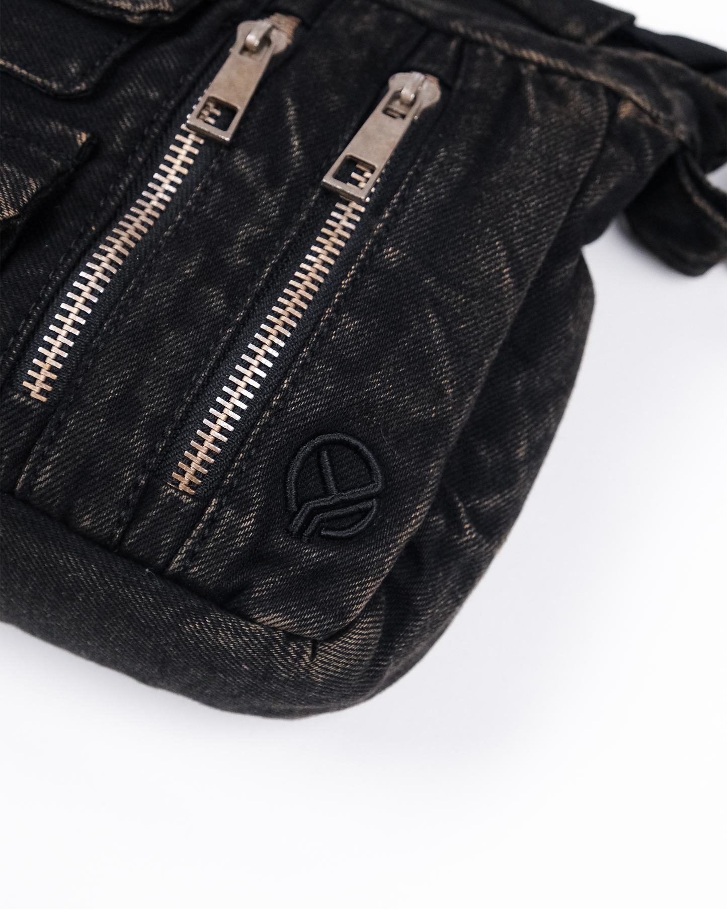 Nahaufnahme einer schwarzen Denim-Umhängetasche mit zwei silberfarbenen Reißverschlüssen und einem aufgestickten Logo in Schwarz in der rechten unteren Ecke.