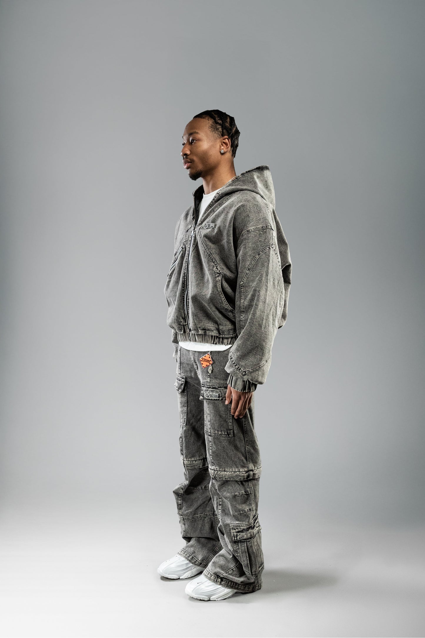 Mann trägt eine hellgraue Jeansjacke mit Kapuze in Seitenansicht. Die Jacke hat breite Ärmel, elastische Bündchen, sichtbare Nähte und eine schräge Reißverschlusstasche. Darunter ist ein weißes Shirt sichtbar. Passend dazu trägt er eine weite Cargo-Jeans aus dem gleichen Denim.