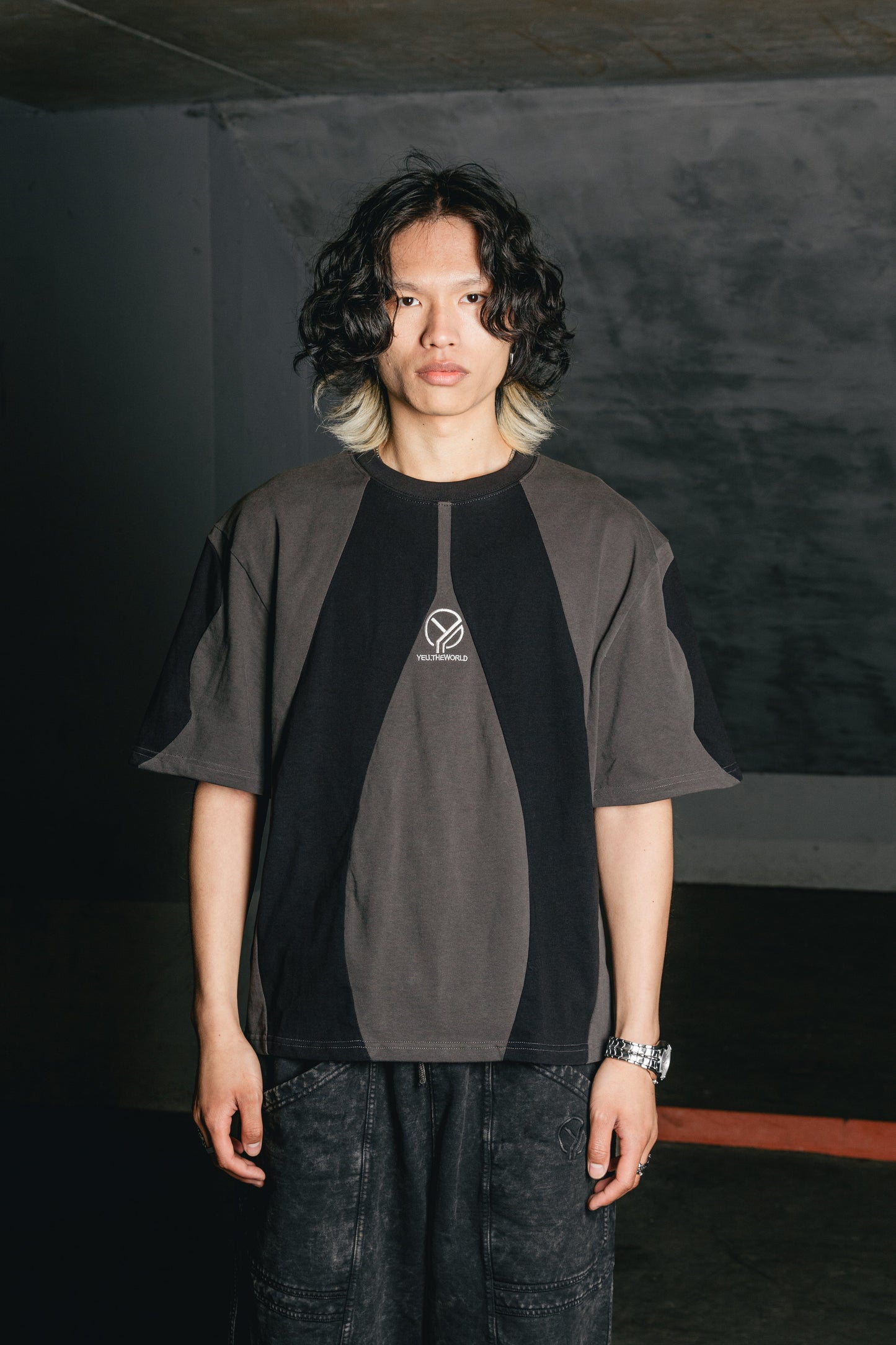 Das schwarze T-Shirt mit grauen, organisch geschwungenen Einsätzen wird von einem männlichen Model in einer neutralen Umgebung getragen. Die Stickerei „YEU.THEWORLD“ ist mittig auf der Brust platziert. Die geometrischen Panels betonen einen futuristischen Look.