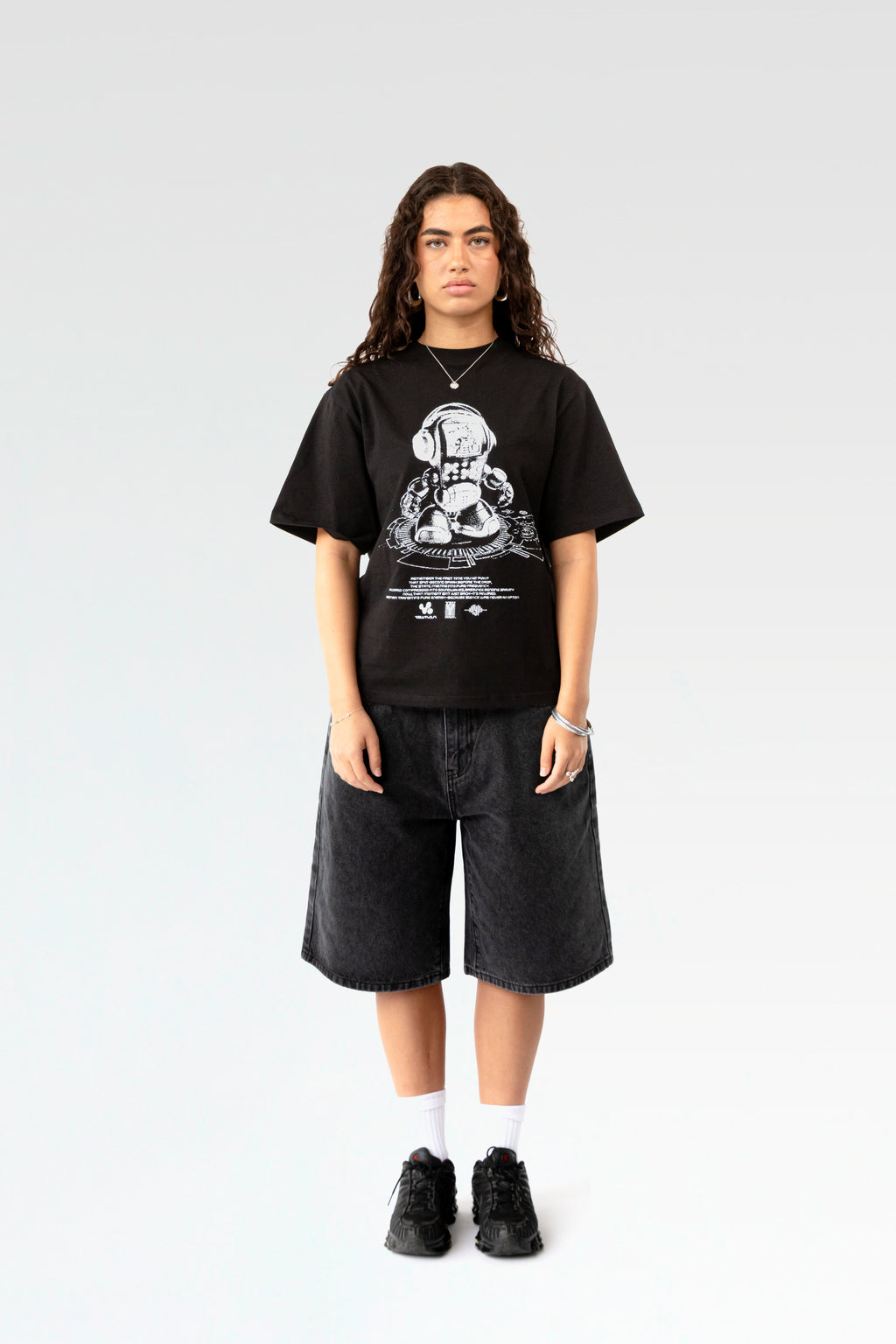 Frontale Studioaufnahme eines schwarzen T-Shirts mit weißem Frontprint. Der Druck zeigt einen stilisierten Roboter mit Kopfhörern auf einem DJ-Pult, darunter Textzeilen und Logos. Das T-Shirt hat einen lockeren Schnitt und wird von einer Person mit dunklen Locken getragen.