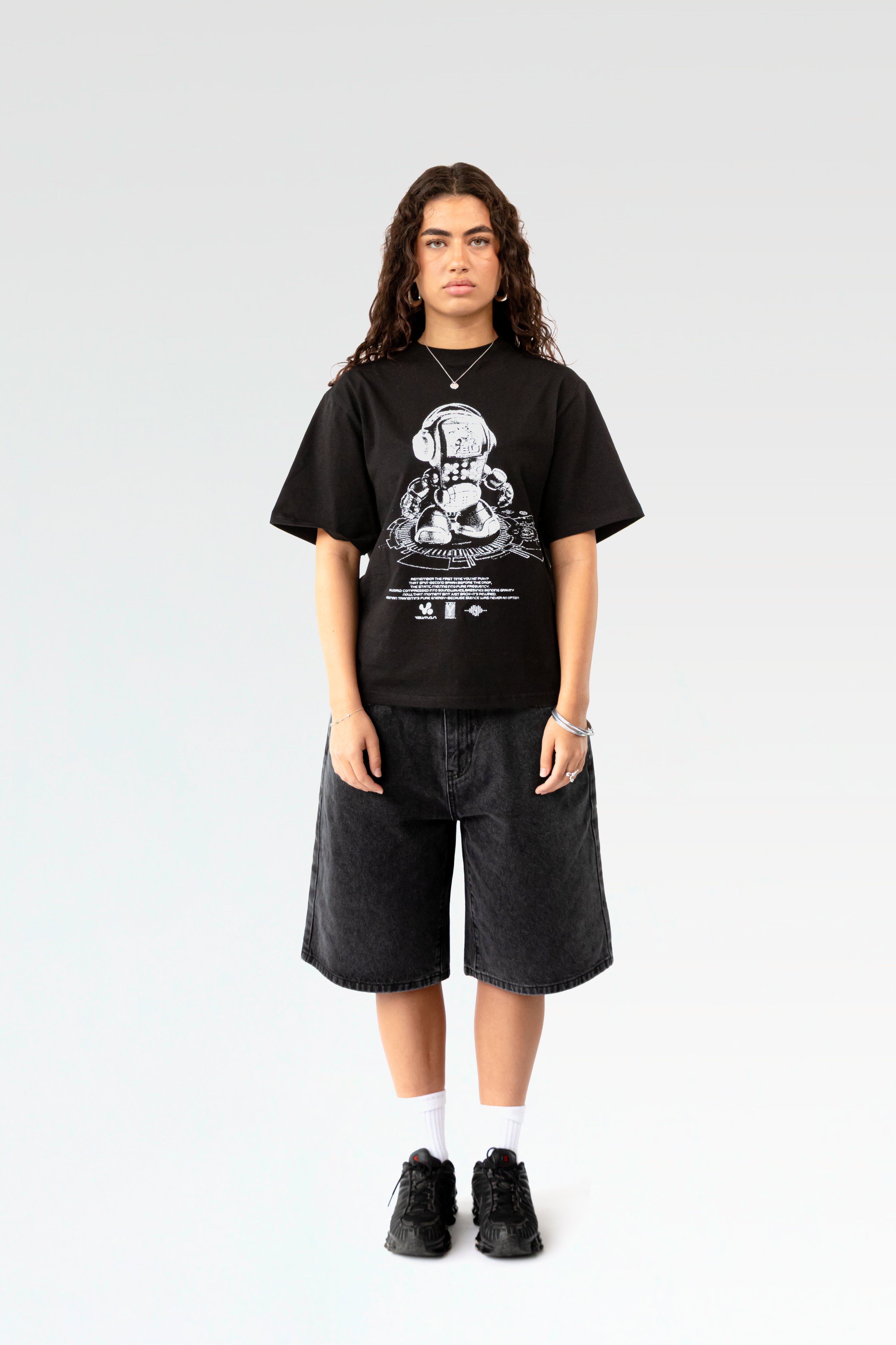 Frontale Studioaufnahme eines schwarzen T-Shirts mit weißem Frontprint. Der Druck zeigt einen stilisierten Roboter mit Kopfhörern auf einem DJ-Pult, darunter Textzeilen und Logos. Das T-Shirt hat einen lockeren Schnitt und wird von einer Person mit dunklen Locken getragen.