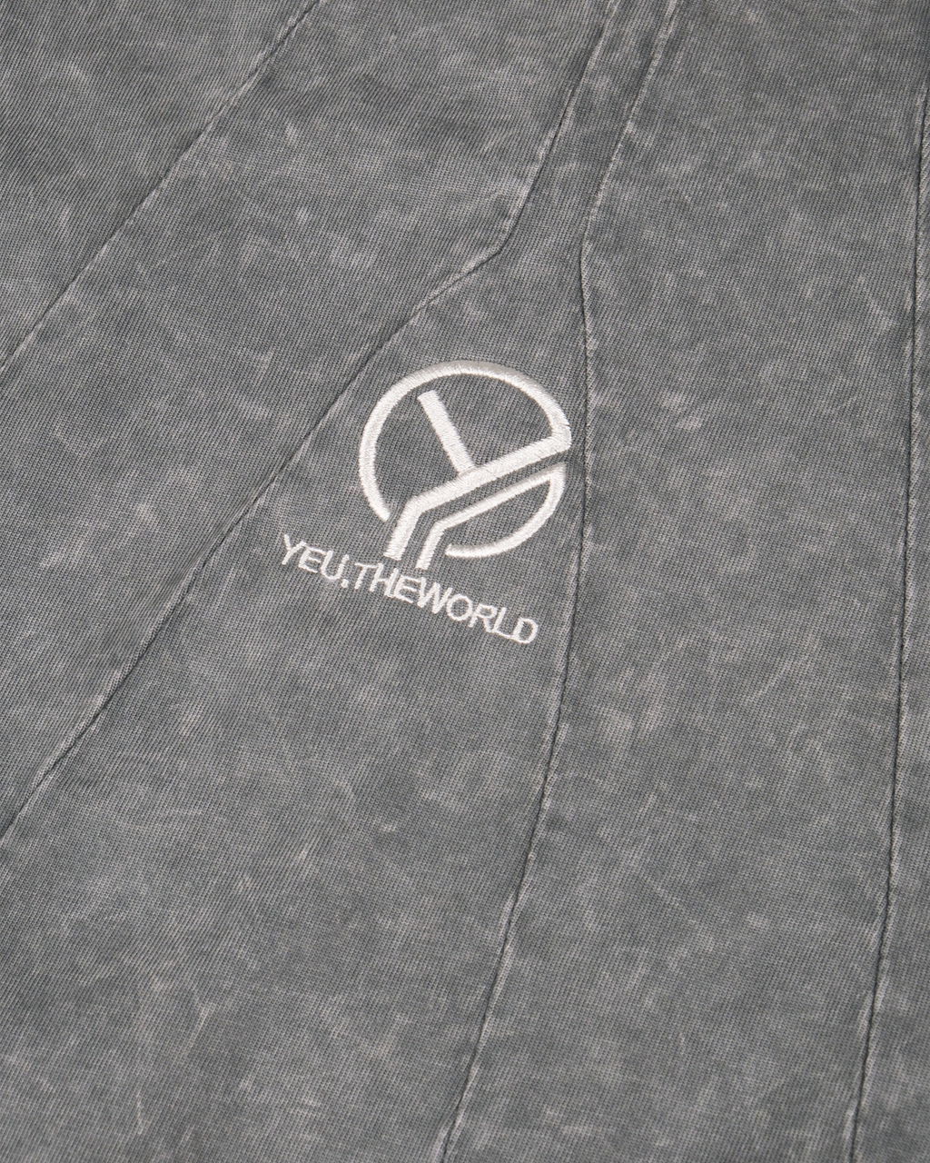 Nahaufnahme eines grauen Langarmshirts mit verwaschener Struktur. Im Brustbereich ist das weiße gestickte Logo „YEU.THEWORLD“ samt stilisiertem Y-Symbol auf einem geometrisch eingepassten Stoffpanel zu sehen.
