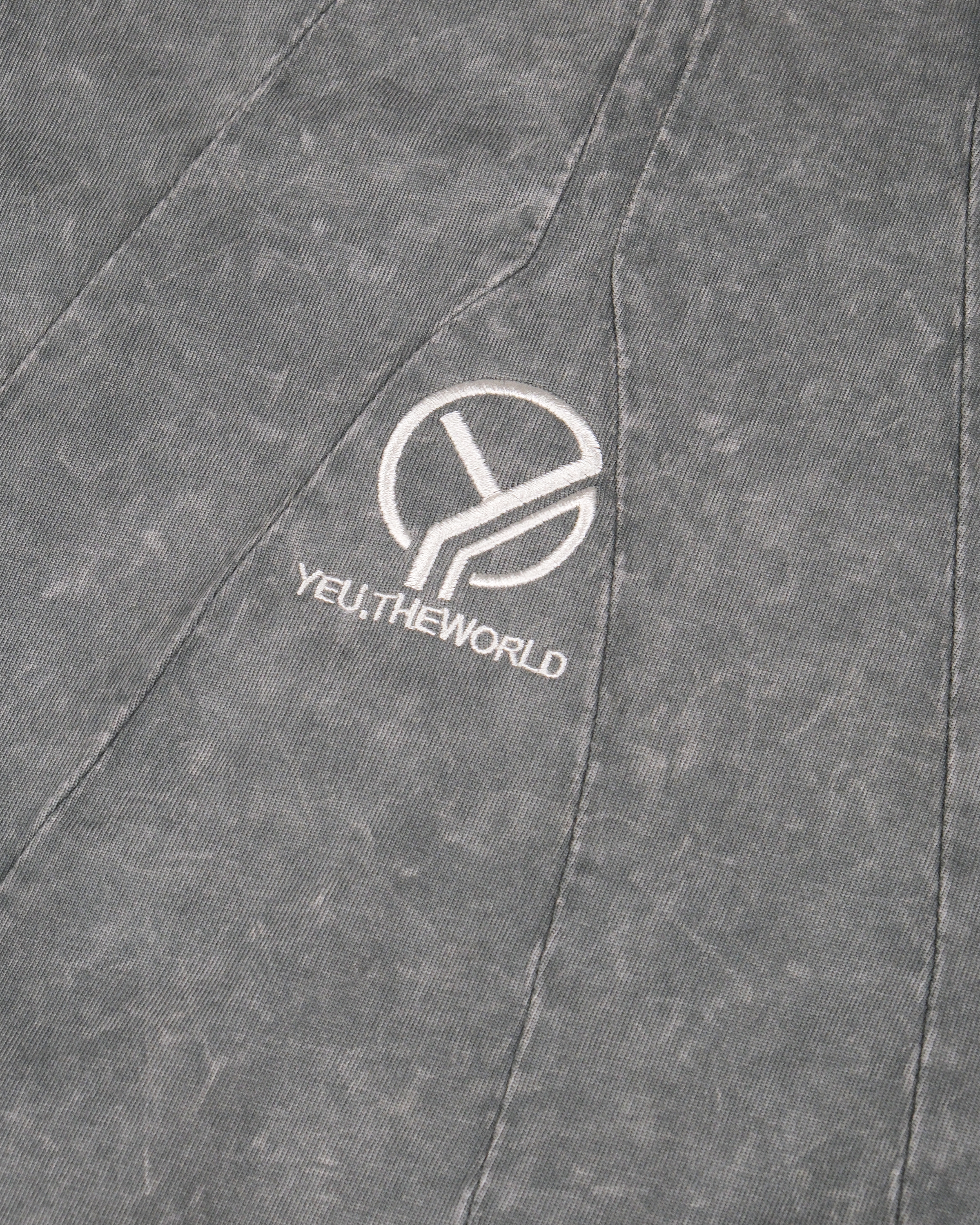 Nahaufnahme eines grauen Langarmshirts mit verwaschener Struktur. Im Brustbereich ist das weiße gestickte Logo „YEU.THEWORLD“ samt stilisiertem Y-Symbol auf einem geometrisch eingepassten Stoffpanel zu sehen.