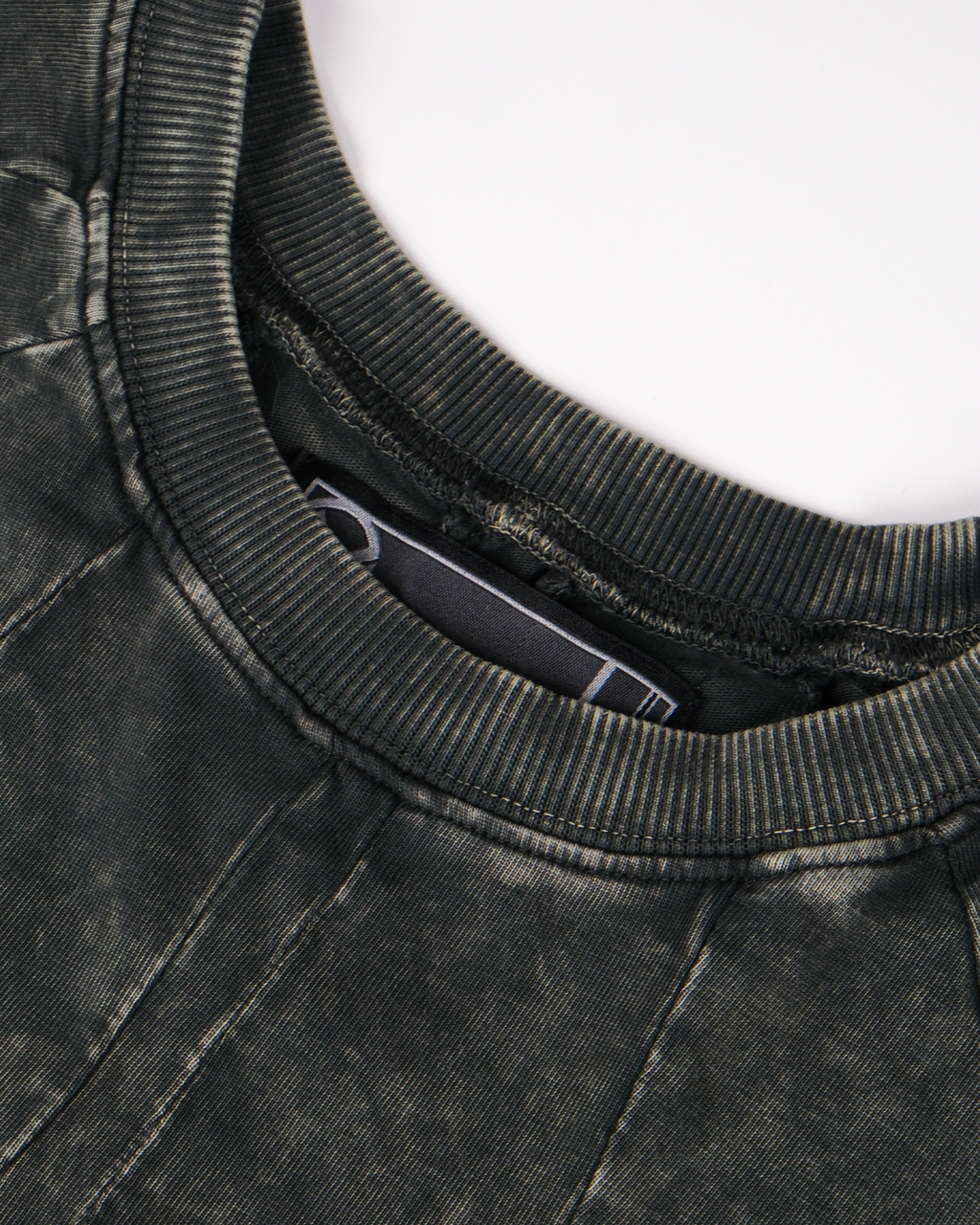  Detailansicht des Halsausschnitts des Shirts. Der Rippenkragen ist in verwaschenem Dunkelgrün gehalten, die umliegende Naht ist sichtbar verarbeitet, im Innern ist ein schwarzes Label mit weißer grafischer Linie zu erkennen.