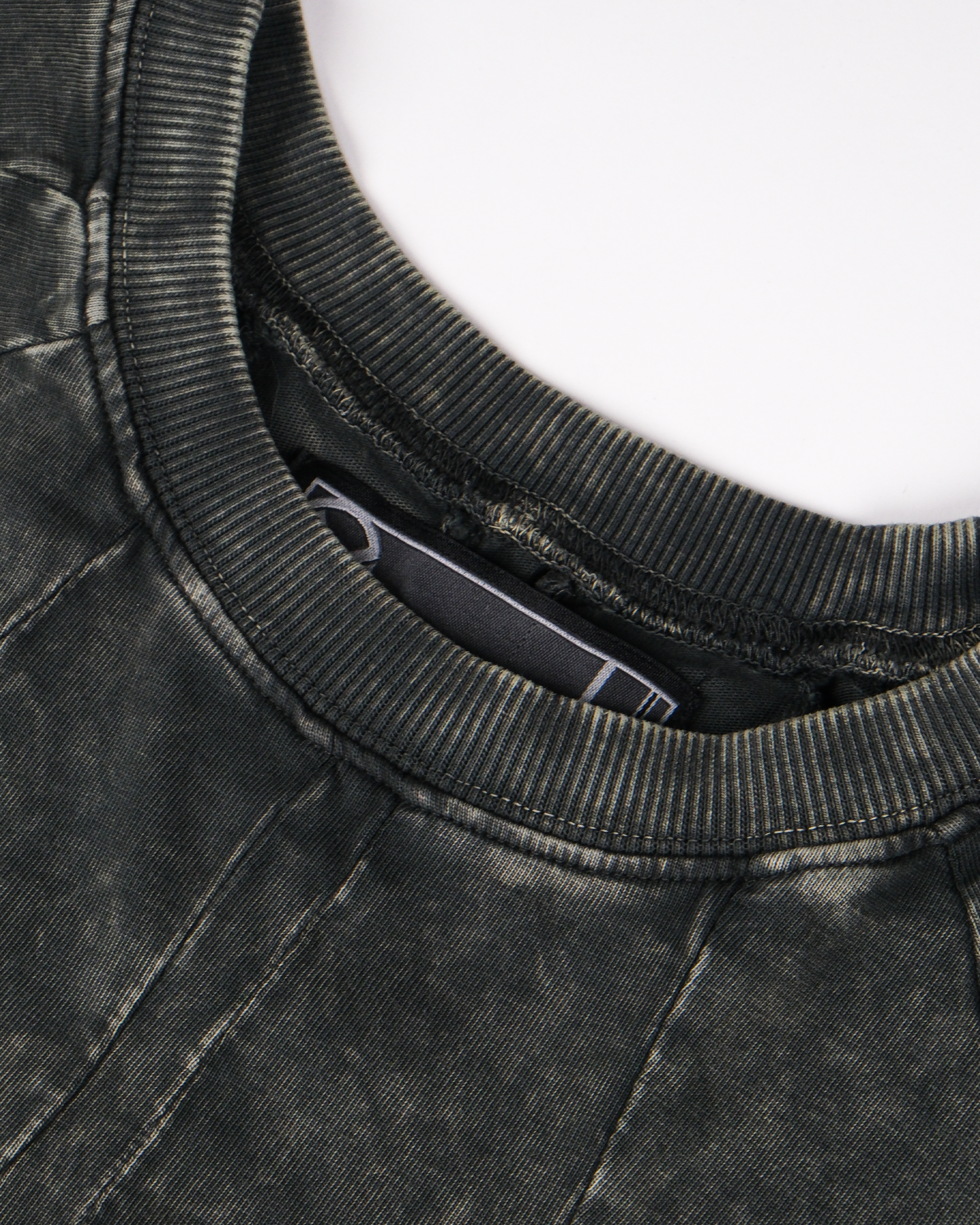  Detailansicht des Halsausschnitts des Shirts. Der Rippenkragen ist in verwaschenem Dunkelgrün gehalten, die umliegende Naht ist sichtbar verarbeitet, im Innern ist ein schwarzes Label mit weißer grafischer Linie zu erkennen.