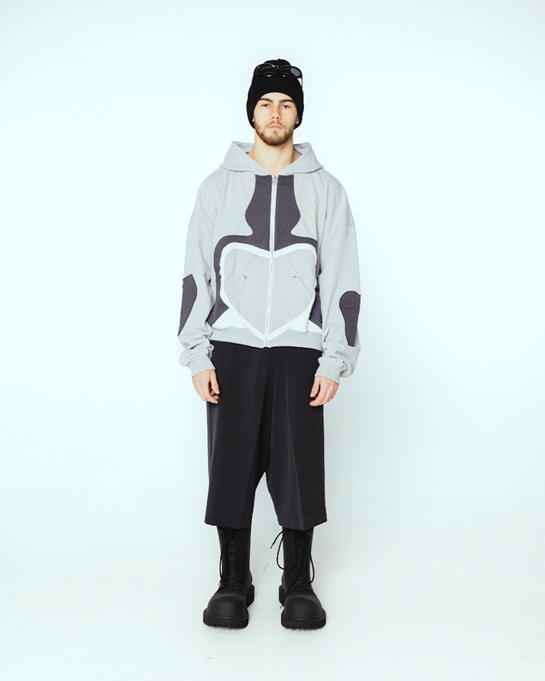 Grauer Zip-Hoodie mit schwarzem Herzmotiv auf der Vorderseite, weißen Kontrastnähten und seitlichen Eingrifftaschen mit Druckknöpfen, getragen von einer Person mit Zöpfen und Sonnenbrille.