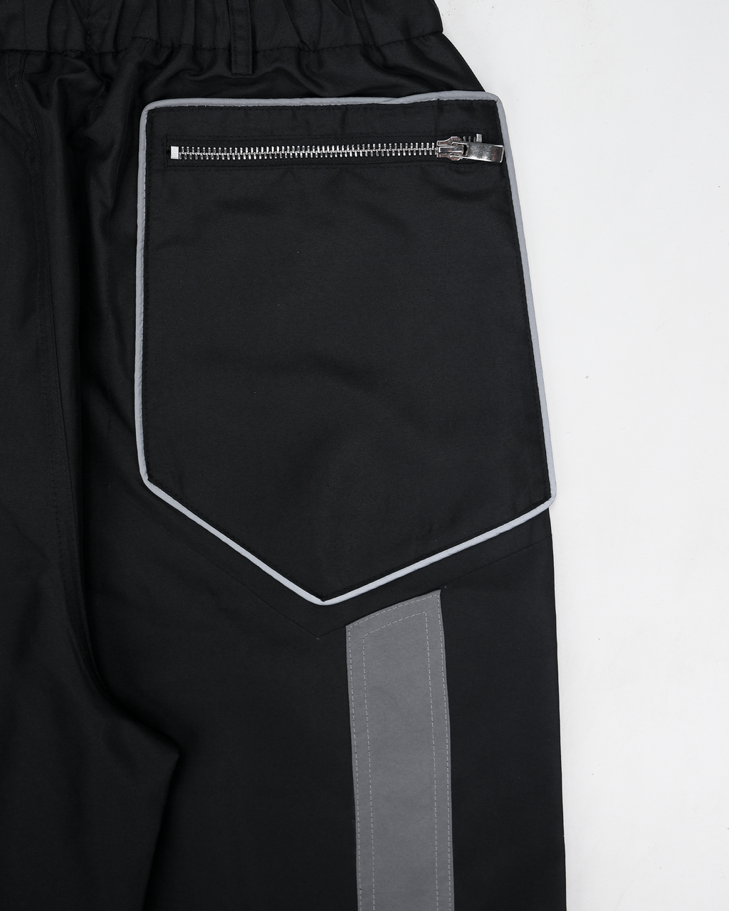 Nahaufnahme der Gesäßtasche einer schwarzen Hose mit technischem Design. Die aufgesetzte Tasche ist fünfeckig geformt und mit einer hellgrauen Paspel eingefasst. Oben verläuft ein horizontaler, silberner Reißverschluss mit glänzendem Zipper. Unterhalb der Tasche beginnt ein grauer, vertikal verlaufender Stoffeinsatz mit sichtbarer Kontrastnaht. Der Bund ist elastisch gerafft.









