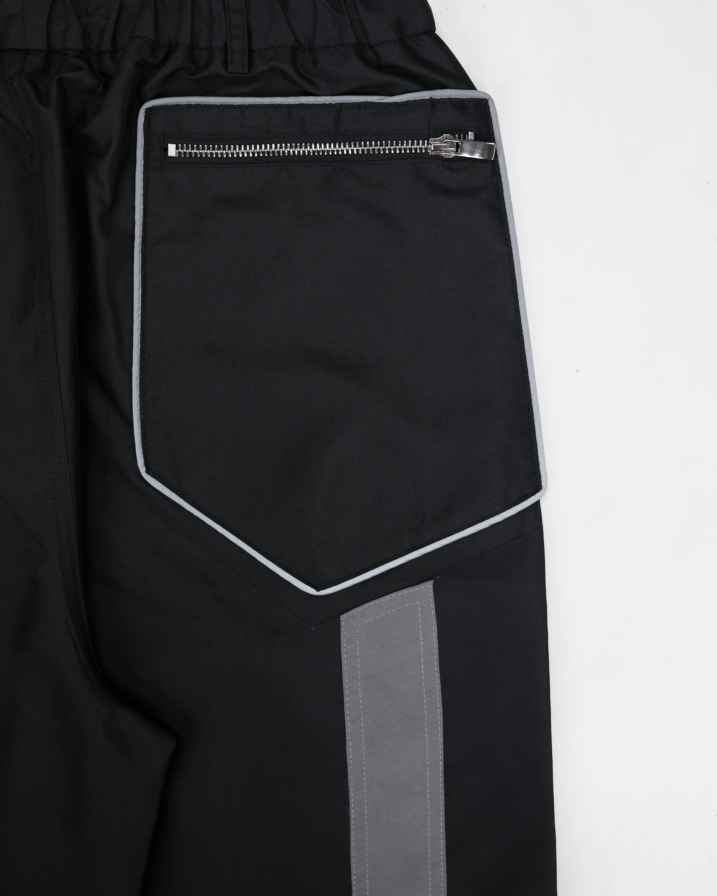 Nahaufnahme der Gesäßtasche einer schwarzen Hose mit technischem Design. Die aufgesetzte Tasche ist fünfeckig geformt und mit einer hellgrauen Paspel eingefasst. Oben verläuft ein horizontaler, silberner Reißverschluss mit glänzendem Zipper. Unterhalb der Tasche beginnt ein grauer, vertikal verlaufender Stoffeinsatz mit sichtbarer Kontrastnaht. Der Bund ist elastisch gerafft.









