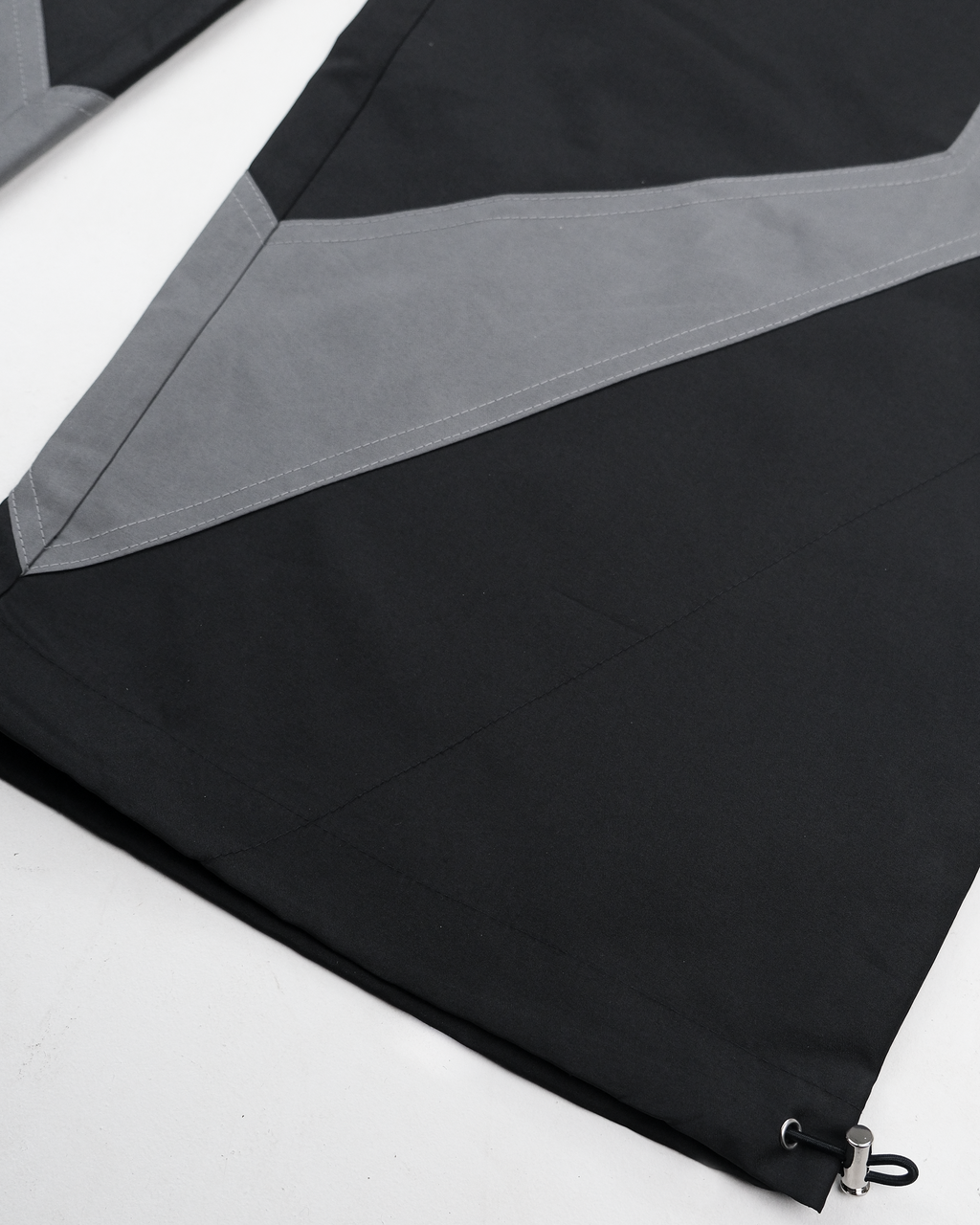 Nahaufnahme des rechten Hosenbeins mit auffälligem, grauem V-förmigem Kontrasteinsatz, der sich mittig nach unten verjüngt. Der schwarze Beinabschluss ist mit einem Tunnelzug ausgestattet, in dem ein schwarzes Gummiband mit silbernem Stopper verläuft. Die saubere Nahtführung unterstreicht die hochwertige Verarbeitung.