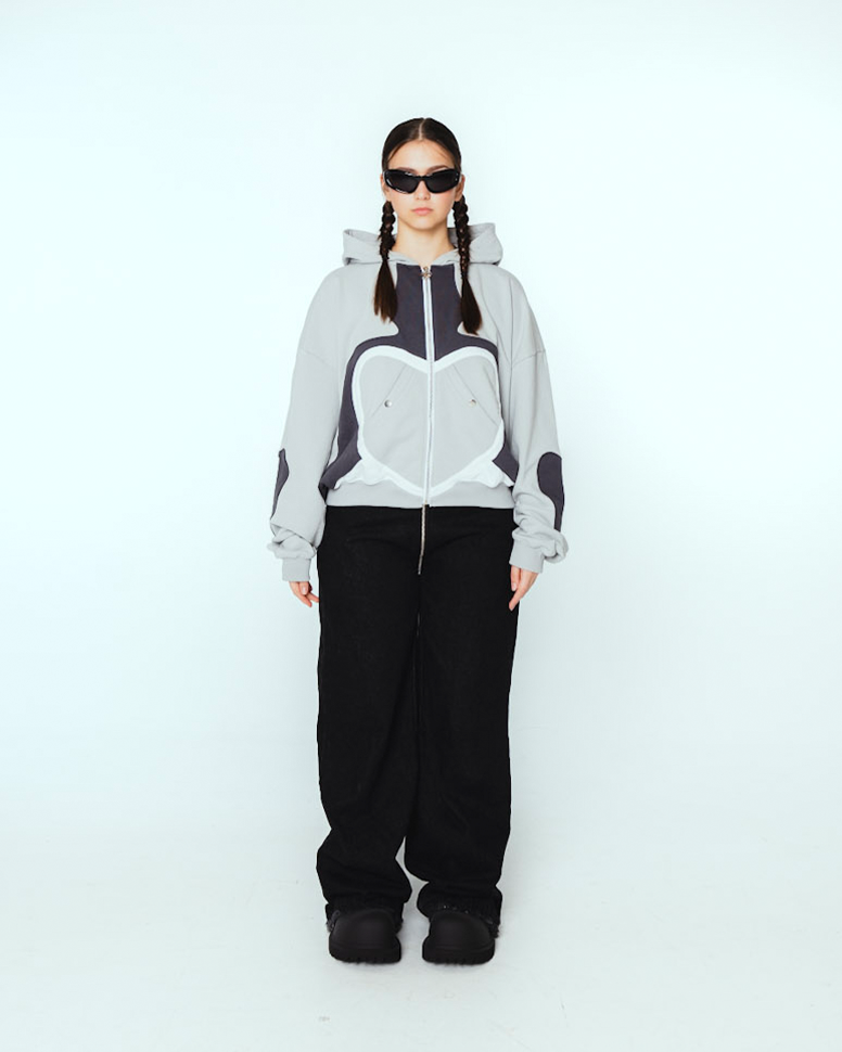 Hellgrauer Zip-Hoodie mit großflächigem Herzmotiv auf der Vorderseite, das aus schwarzen und weißen Stoffapplikationen gebildet ist. Der Reißverschluss verläuft mittig durch das Herzdesign, zwei silberne Druckknöpfe fixieren die darüberliegenden, diagonal eingesetzten Taschen. Die Kapuze liegt flach an, die Ärmel zeigen schwarze Herzapplikationen auf den Außenseiten.