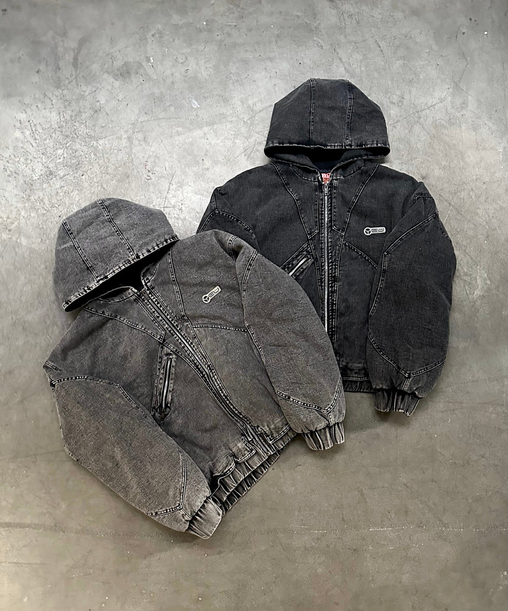 Zwei Denim-Kapuzenjacken in gewaschenem Schwarz und Grau liegen flach nebeneinander auf Betonboden. Beide Modelle haben einen Reißverschluss, schräge Eingrifftaschen und ein kleines Logo-Patch auf der linken Brust.