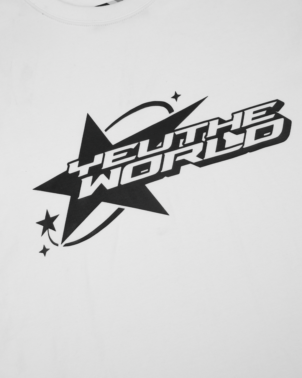 Nahaufnahme des Frontprints auf weißem Stoff. Schwarze Illustration bestehend aus einem großen Stern mit drei kleinen Sternen links unten. Zentraler Schriftzug: „YEU THE WORLD“ in kastenförmiger Typografie mit Bogenlinie und Glanzeffekt-Optik.