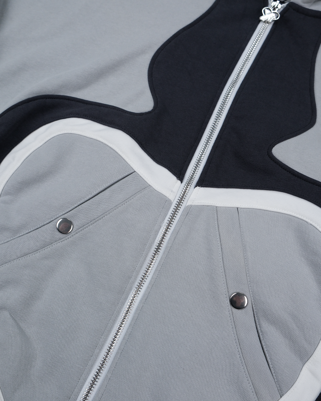 Nahaufnahme der seitlichen Eingrifftasche mit Druckknopf auf dem grauen Hoodie. Sichtbar sind die schwarze Taschenklappe und die weiße Kontrastpaspelierung auf dem strukturierten Stoff.