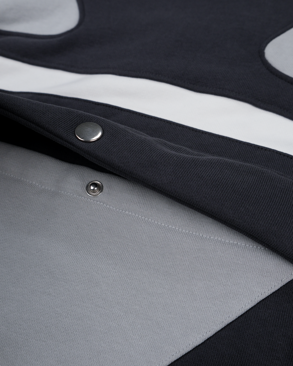 Nahaufnahme der seitlichen Eingrifftasche mit Druckknopf auf dem grauen Hoodie. Sichtbar sind die schwarze Taschenklappe und die weiße Kontrastpaspelierung auf dem strukturierten Stoff.

