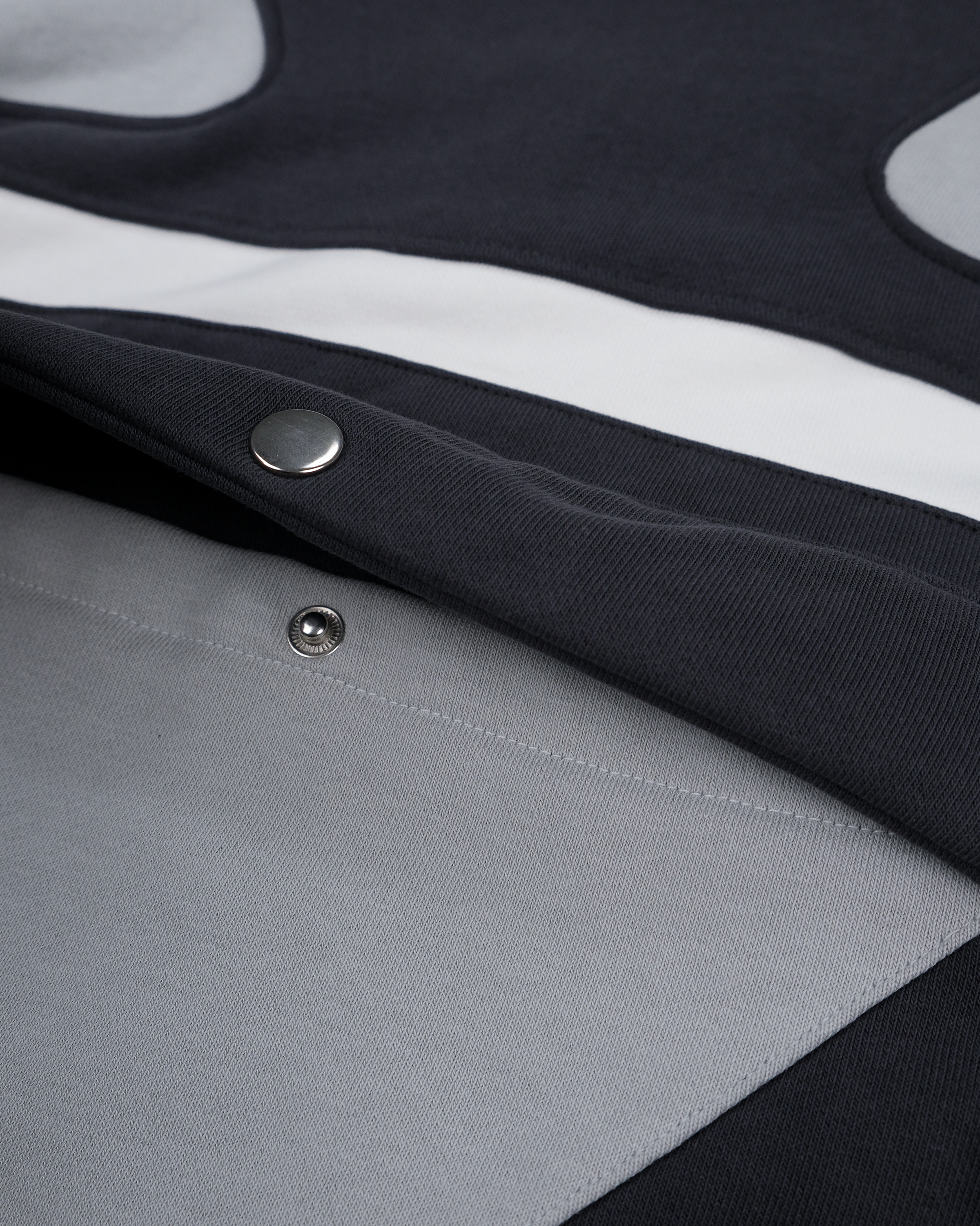 Nahaufnahme der seitlichen Eingrifftasche mit Druckknopf auf dem grauen Hoodie. Sichtbar sind die schwarze Taschenklappe und die weiße Kontrastpaspelierung auf dem strukturierten Stoff.

