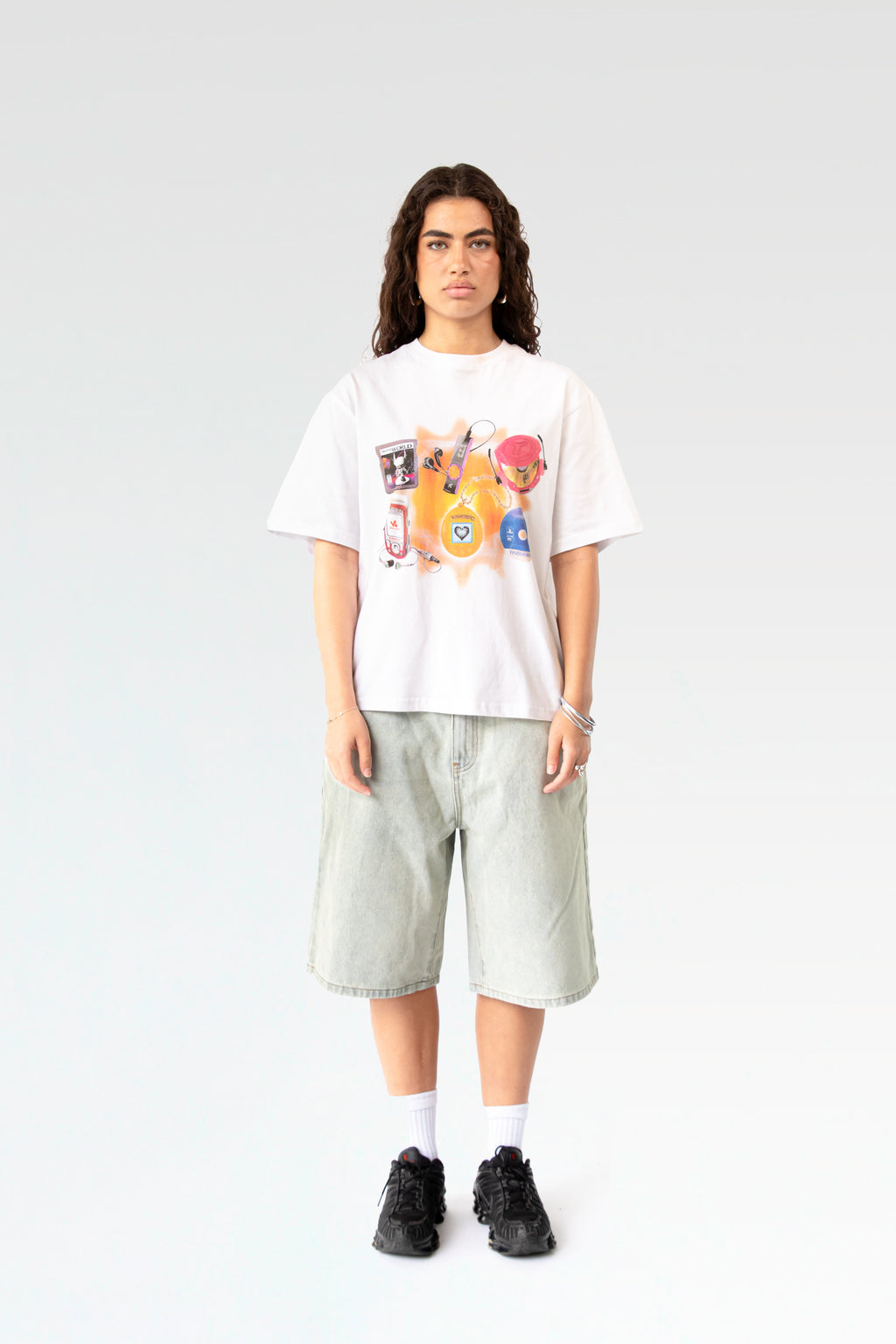 Frontale Studioaufnahme eines weißen T-Shirts mit buntem Frontprint, getragen von einer stehenden Person. Der Druck zeigt Kindheitsobjekte wie MP3-Player, Tamagotchi, Jojo, Spielkassette und Handy auf orangefarbenem Hintergrund.