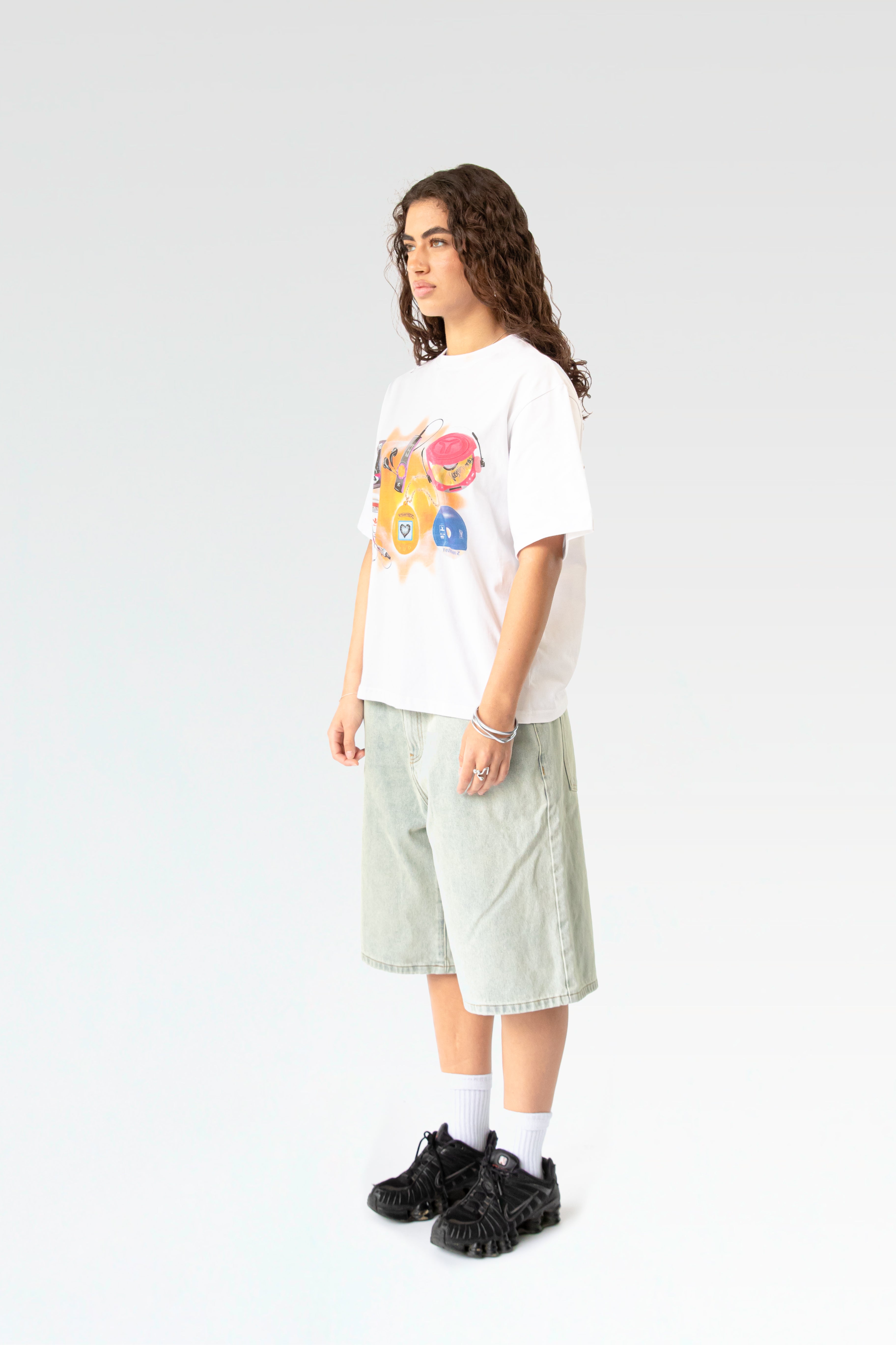 Schräg seitliche Studioaufnahme des weißen T-Shirts mit Kindheits-Print. Das locker geschnittene Shirt wird zu einer hell verwaschenen Denim-Bermuda getragen, die weite Passform ist erkennbar.