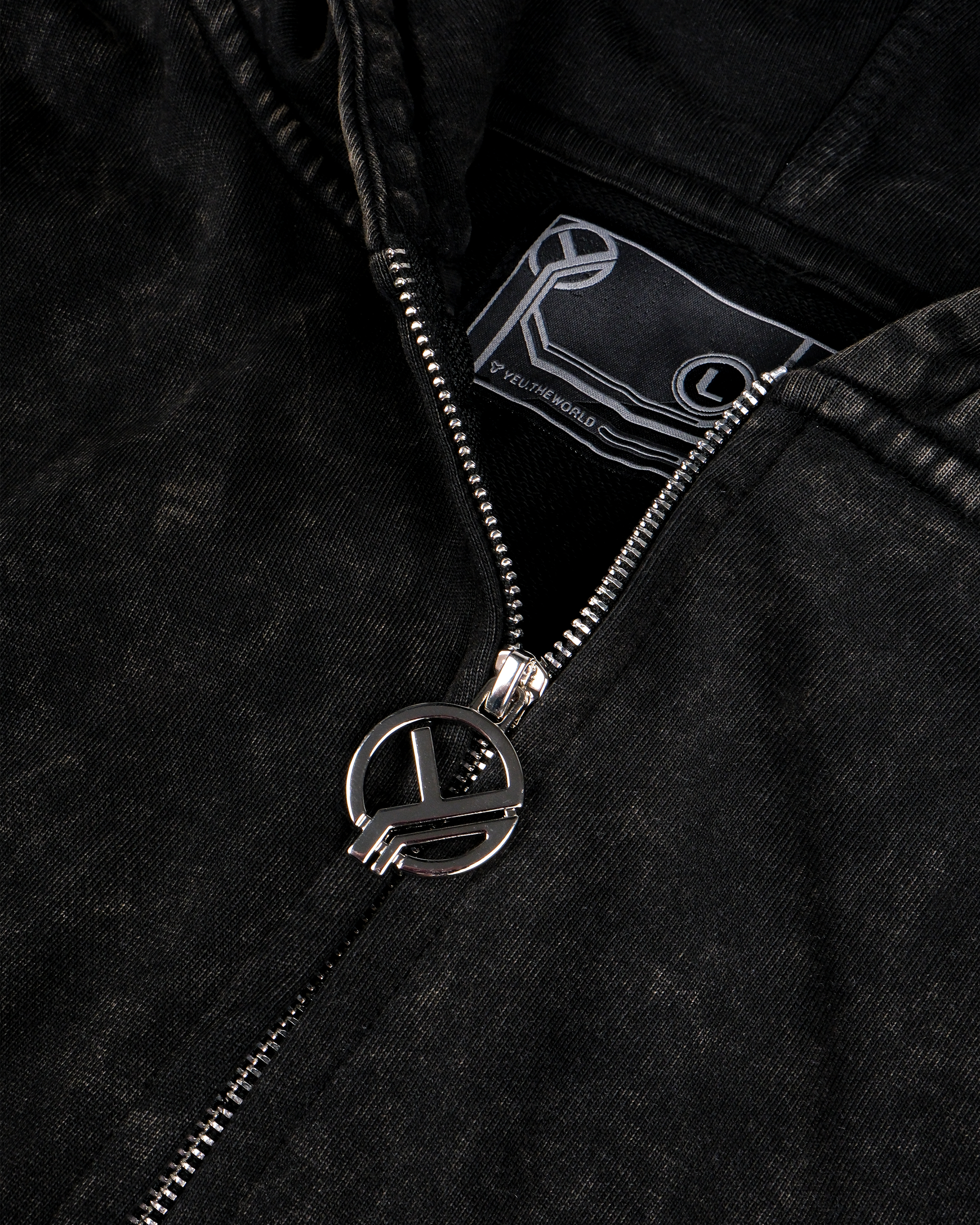 Detailansicht eines schwarzen Zip-Hoodies mit gewaschenem Vintage-Effekt. Gezeigt wird der geöffnete Reißverschluss mit silbernem Zipper in Kreisform, durchkreuzt von einem stilisierten „Y“-Symbol. Im Hintergrund sichtbar ist das gewebte Label mit „YEU THE WORLD“-Schriftzug und Größenangabe „L“.