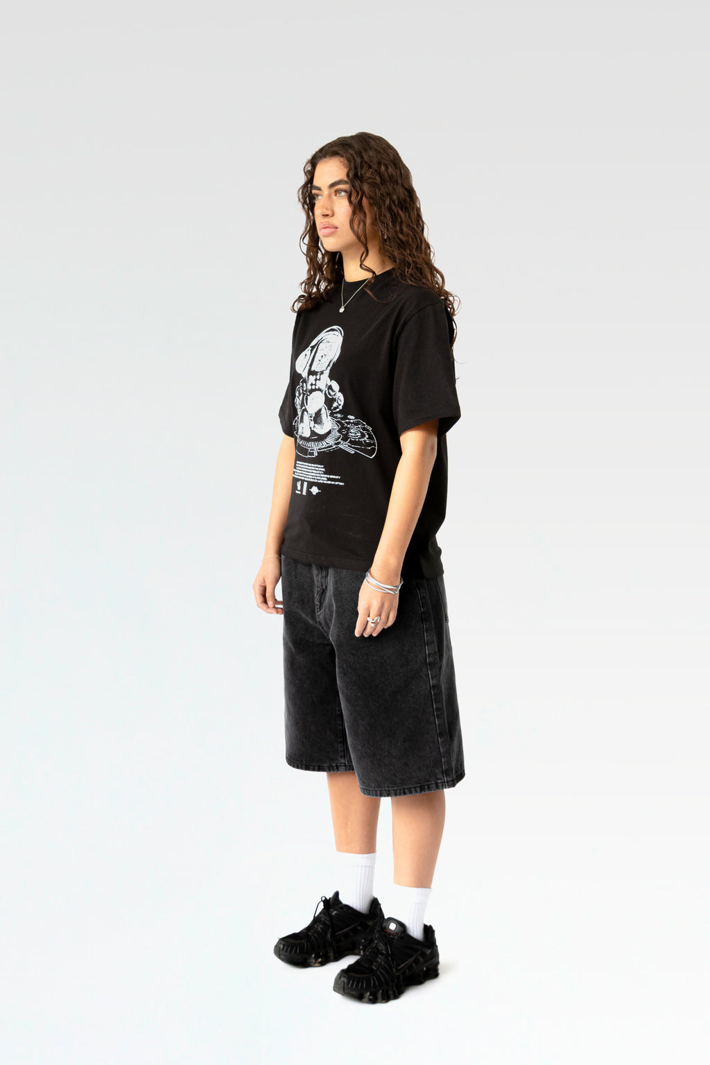 Schräg seitliche Ansicht des schwarzen T-Shirts mit auffälligem Roboter-Motiv. Der weiße Print erstreckt sich mittig über die Brust. Getragen wird das locker geschnittene Shirt über einer hellgrau verwaschenen Jeansshorts.
