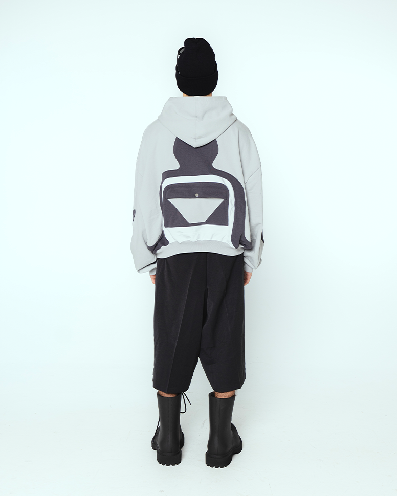 Grauer Kapuzenpullover mit schwarzen und weißen geometrischen Applikationen auf dem Rücken, getragen von einer Person mit schwarzer Mütze und weiten Shorts. Das Rückendesign erinnert an eine stilisierte Rückenform mit mittiger Klappentasche mit Druckknopf.

