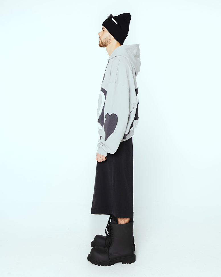 Grauer Kapuzenpullover mit seitlich appliziertem Herz in Schwarz auf dem Ärmel, getragen von einer Person in weitem schwarzen Rock und Gummistiefeln. Die Herzapplikation hebt sich kontrastreich vom Grundton ab.