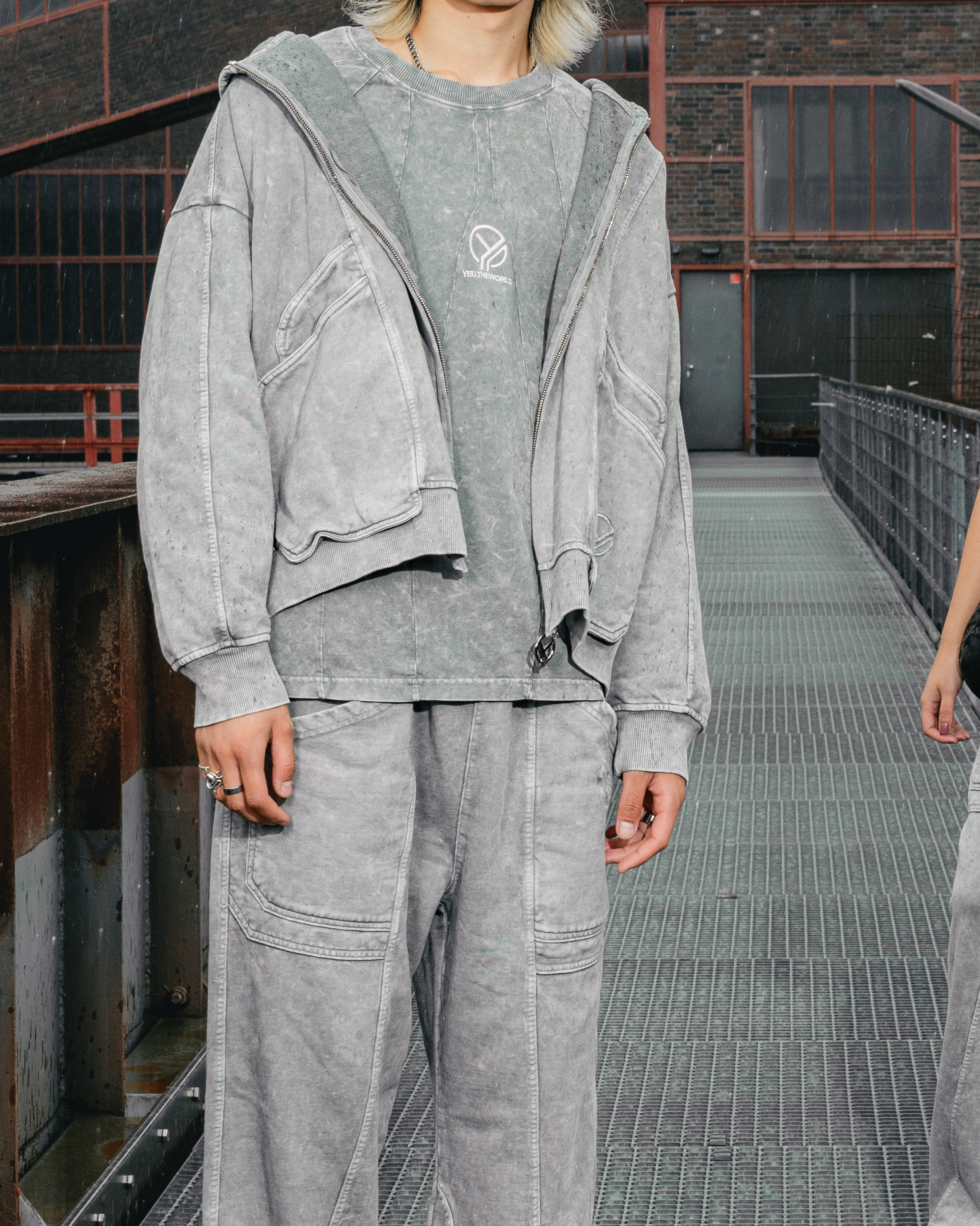 Model trägt ein verwaschen graues Langarmshirt mit gesticktem YEU.THEWORLD-Logo unter einer offenen Zip-Jacke im gleichen verwaschenen Look. Dazu kombiniert ist eine passende, locker geschnittene Cargohose in hellem Grau. Der Fokus liegt auf dem Shirt.