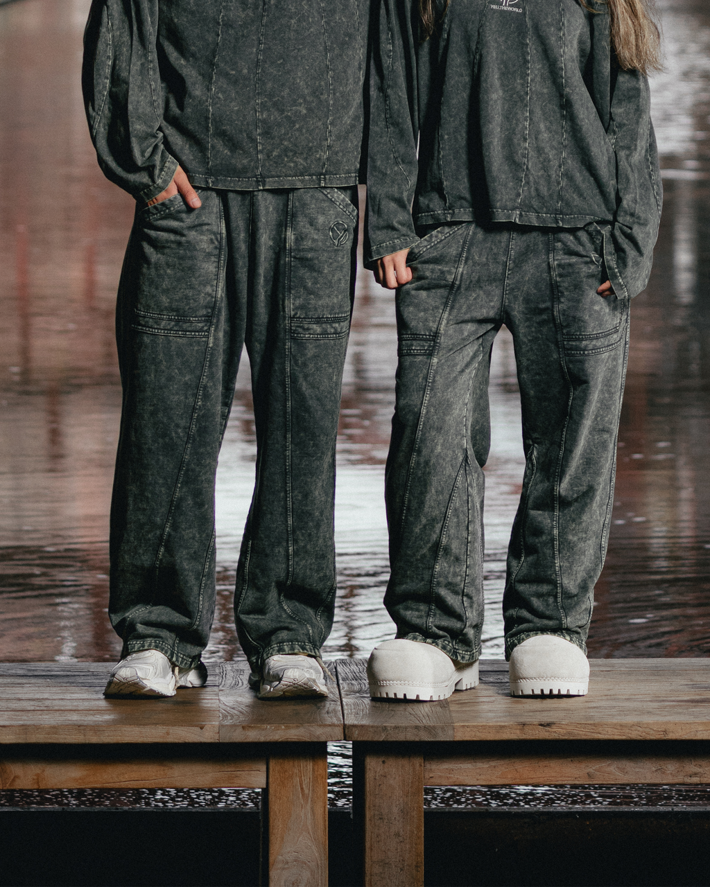 Zwei Personen tragen graue Sweatpants mit ausgewaschener Optik und sichtbaren Teilungsnähten. Die Hosen zeigen schräge Gesäßtaschen und aufgesticktes YEU-Logo oberhalb der rechten Tasche. Beide stehen nebeneinander auf einem Holzsteg vor nassem Untergrund – eine Person trägt klobige weiße Schuhe, die andere Sneaker.