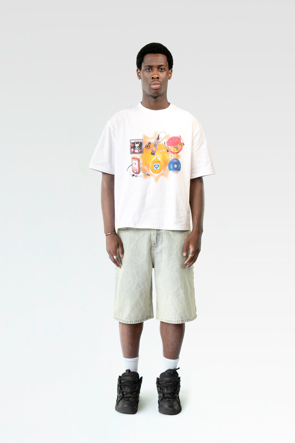 Frontale Studioaufnahme des weißen T-Shirts mit Kindheitsmotiv, getragen von einer männlich gelesenen Person. Der bunte Print hebt sich vom weißen Stoff ab, die Hose ist hell verwaschen.