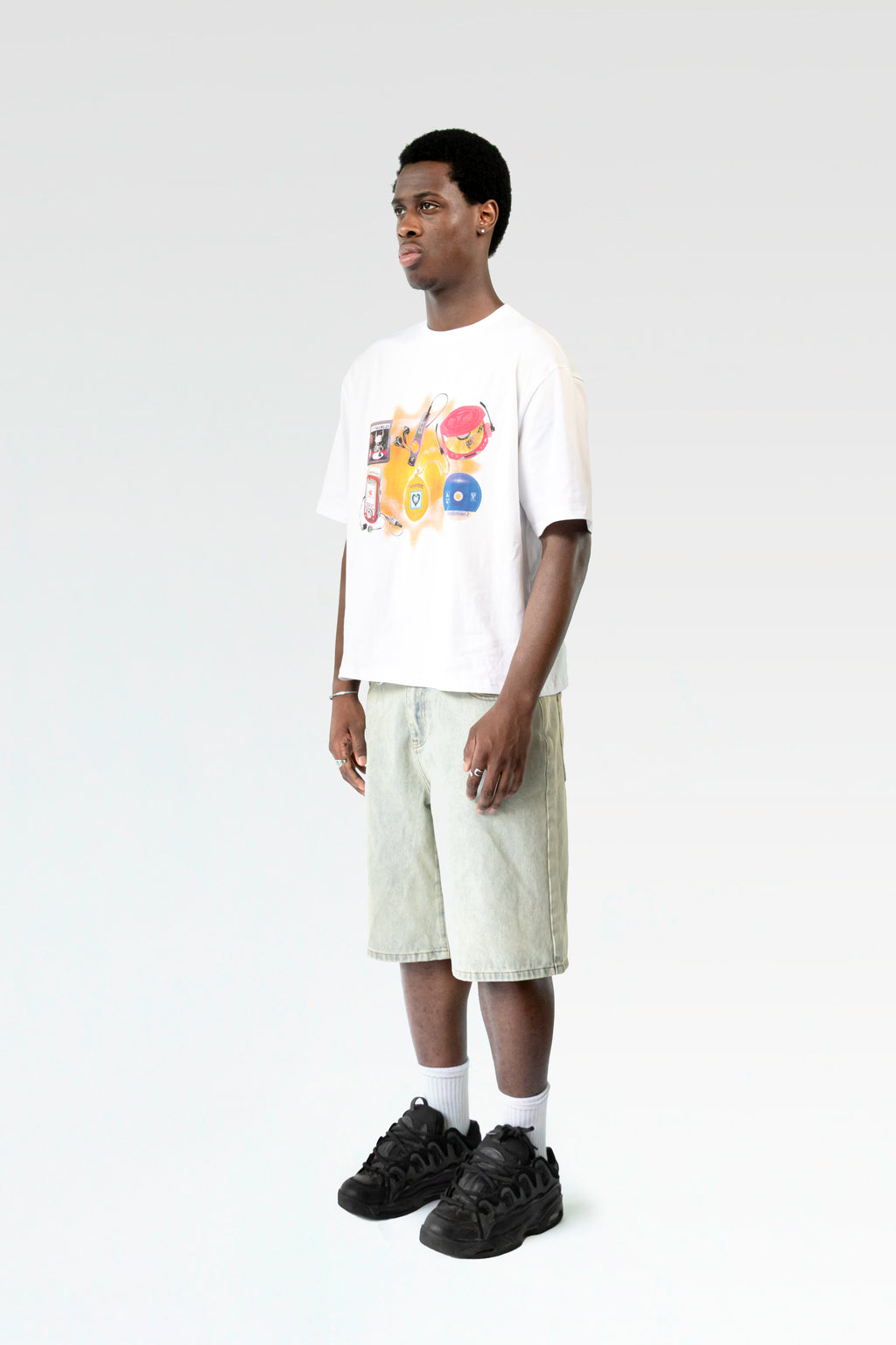 Schräg seitliche Studioaufnahme des weißen T-Shirts mit Childhood-Motiv, getragen von einer männlich gelesenen Person. Das Shirt fällt locker, die Shorts hat eine helle Waschung.