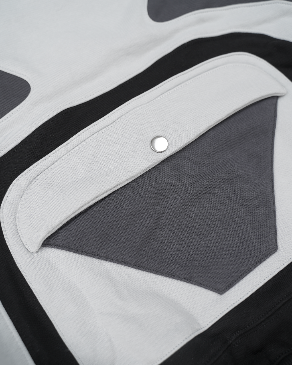Nahaufnahme der Rückseite des Hoodies mit mehrlagiger Applikation. Sichtbar ist eine dunkelgraue, dreieckige Tasche mit darüberliegender, weißer Klappe und silbernem Druckknopf. Die Elemente sind von kontrastierenden Stoffbahnen in Weiß und Schwarz eingefasst, die eine stilisierte Herzform ergeben. Saubere Nähte und präzise Verarbeitung der Materialien.









