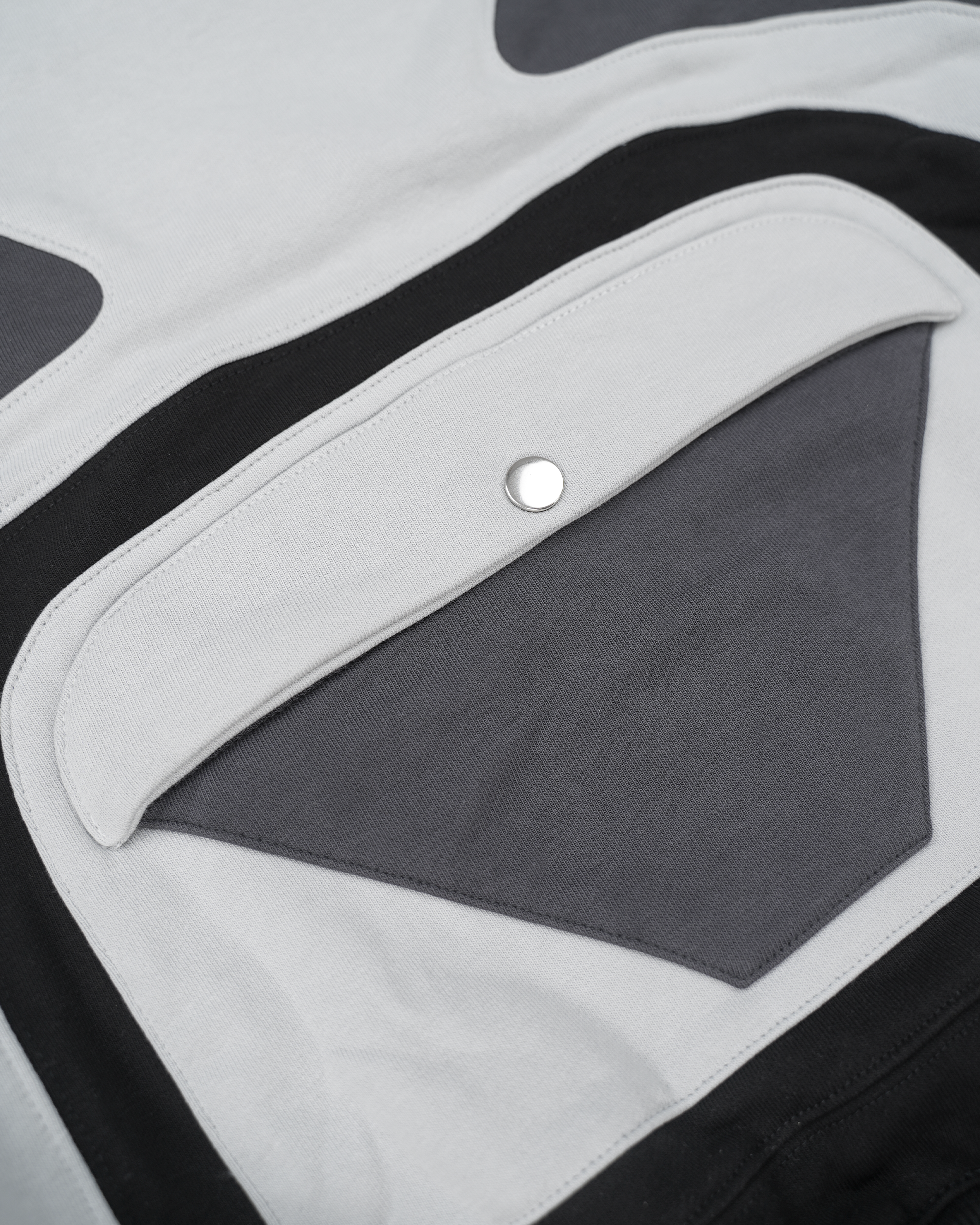 Nahaufnahme der Rückseite des Hoodies mit mehrlagiger Applikation. Sichtbar ist eine dunkelgraue, dreieckige Tasche mit darüberliegender, weißer Klappe und silbernem Druckknopf. Die Elemente sind von kontrastierenden Stoffbahnen in Weiß und Schwarz eingefasst, die eine stilisierte Herzform ergeben. Saubere Nähte und präzise Verarbeitung der Materialien.









