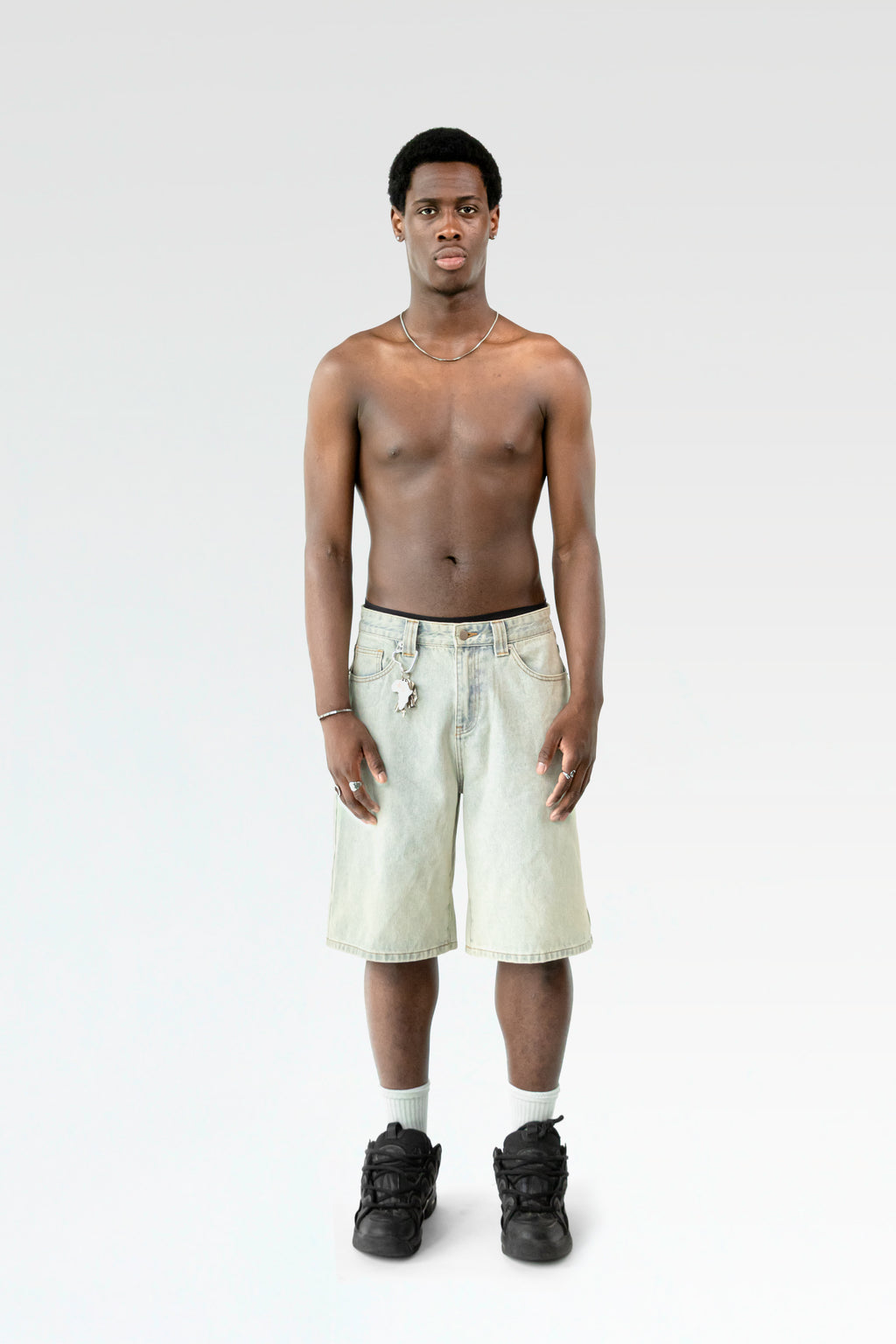 Frontale Studioaufnahme einer hellblauen Bermuda-Shorts an einer männlich gelesenen Person. Die Shorts endet knapp über dem Knie und ist mit einem Karabiner samt Schlüsselbund an der Gürtelschlaufe ausgestattet.