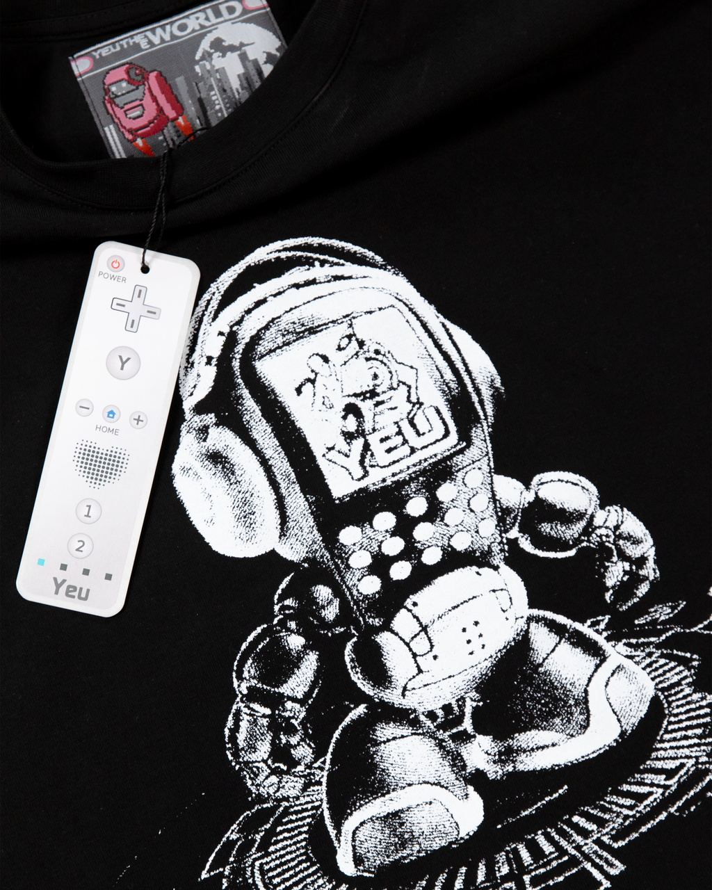 Nahaufnahme des Prints auf dem schwarzen T-Shirt. Der Druck zeigt einen pixelartigen Roboter mit Kopfhörern und DJ-Pult. Vor dem Shirt hängt ein weißer Controller als Merchandise-Anhänger mit der Aufschrift „Yeu“.