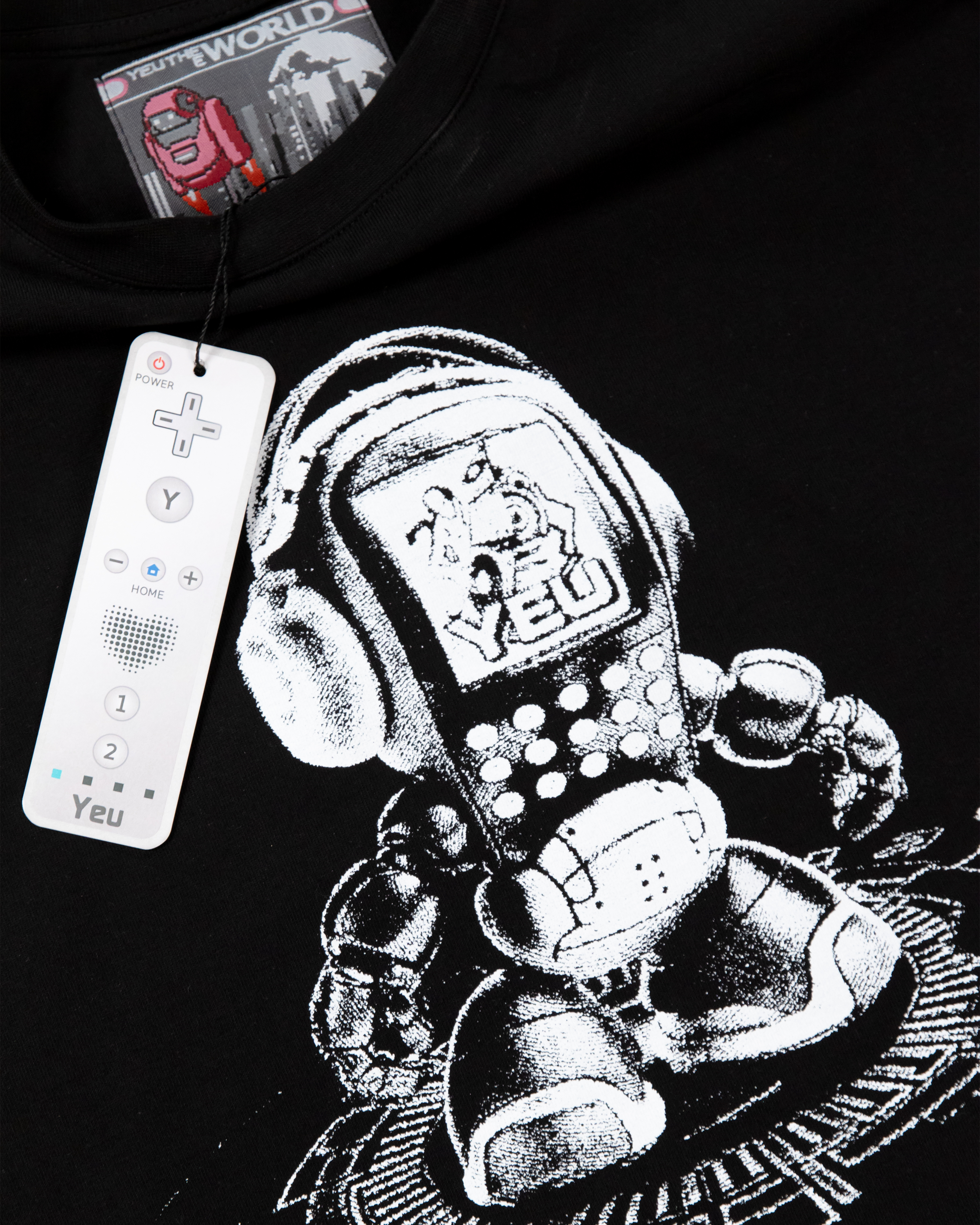 Nahaufnahme des Prints auf dem schwarzen T-Shirt. Der Druck zeigt einen pixelartigen Roboter mit Kopfhörern und DJ-Pult. Vor dem Shirt hängt ein weißer Controller als Merchandise-Anhänger mit der Aufschrift „Yeu“.