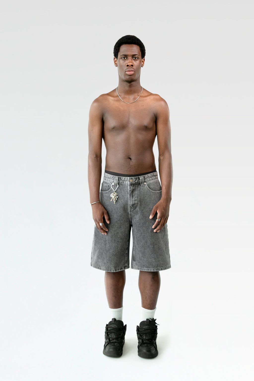Frontale Studioaufnahme einer hellgrau verwaschenen Bermuda-Shorts an einer männlich gelesenen Person. Die Shorts endet knapp über dem Knie und ist mit einem Karabiner samt Schlüsselbund an der Gürtelschlaufe ausgestattet.