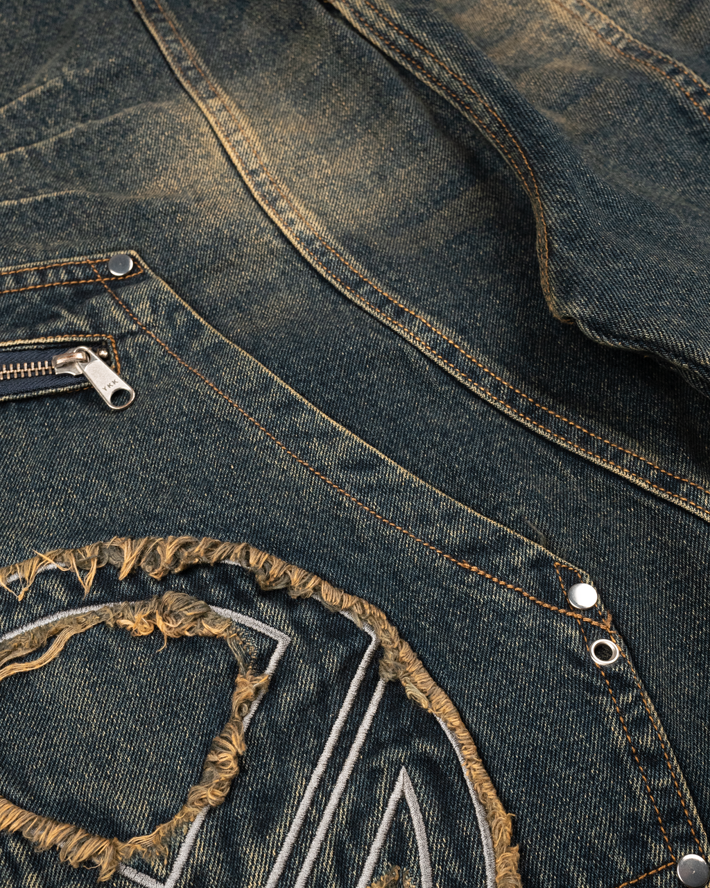 Nahaufnahme der Denim-Shorts mit Fokus auf die ausgefranste Applikation in Form eines Kreises und gesticktem Motiv, dazu sichtbare Kontrastnähte und silberner Reißverschluss der Seitentasche.