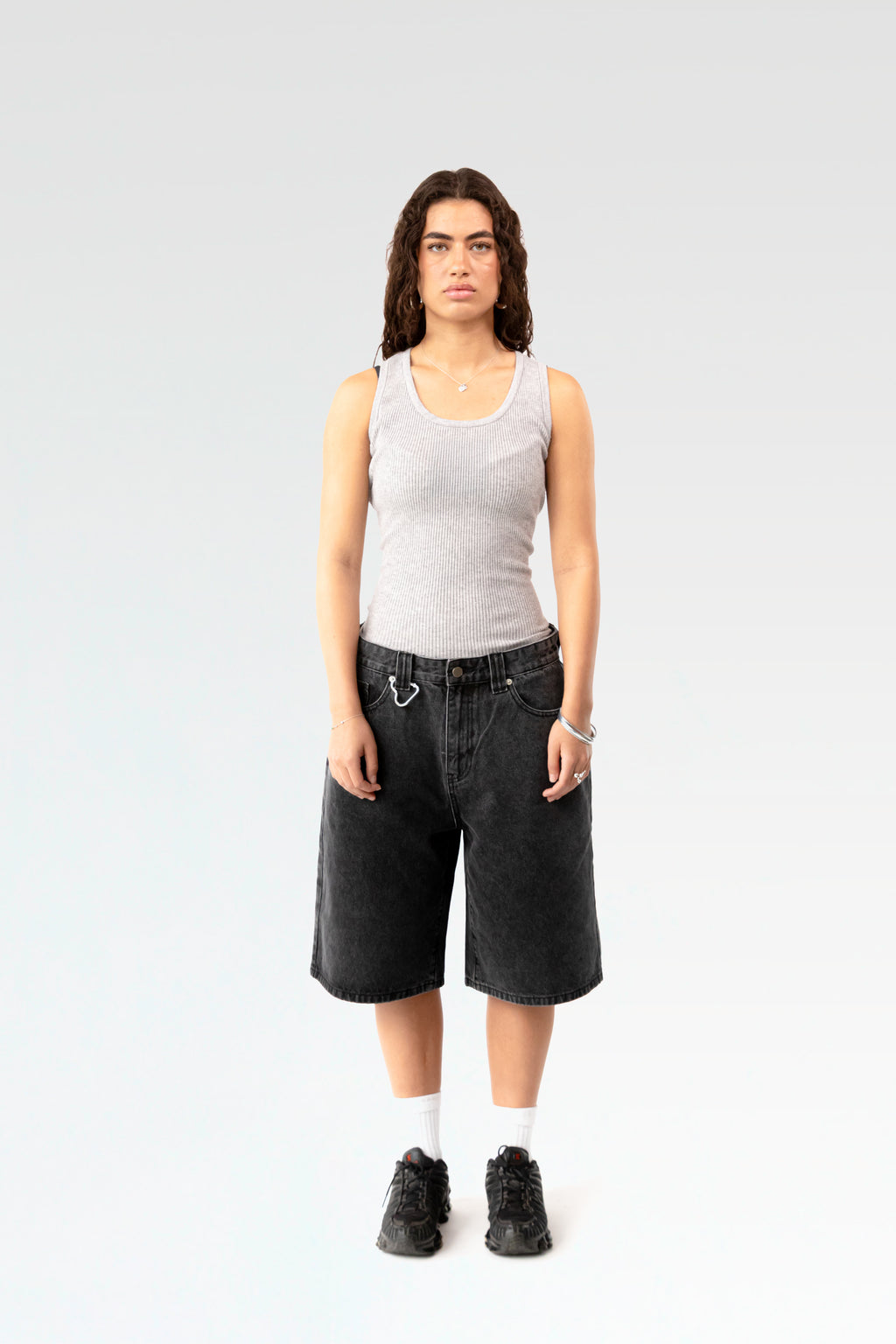 Frontale Studioaufnahme einer schwarzen Denim-Bermuda-Shorts an einer stehenden Person. Die Shorts ist knielang geschnitten und hat eine weite Passform sowie einen silbernen Karabinerhaken an der Gürtelschlaufe.