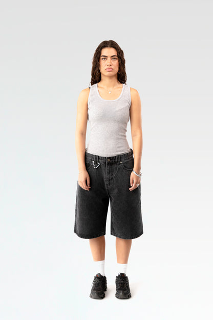 Frontale Studioaufnahme einer schwarzen Denim-Bermuda-Shorts an einer stehenden Person. Die Shorts ist knielang geschnitten und hat eine weite Passform sowie einen silbernen Karabinerhaken an der Gürtelschlaufe.