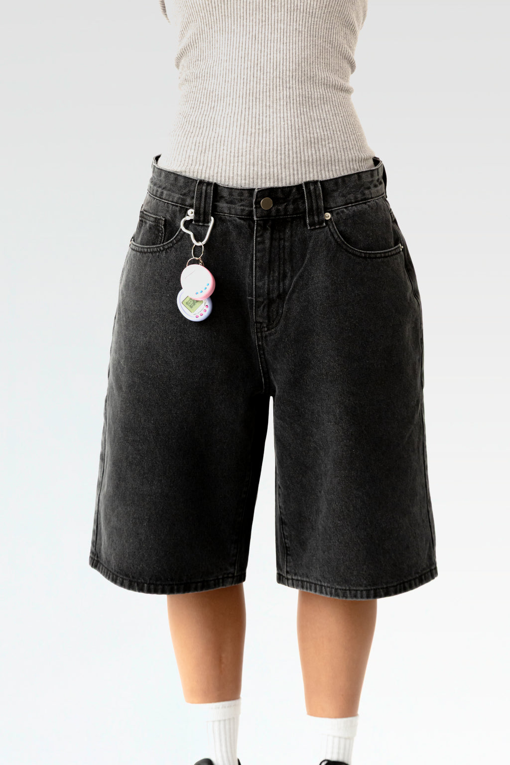 Vorderansicht einer schwarzen, knielangen Jeansshorts aus Waschdenim an einer stehenden Person. Die Shorts hat einen Herz-förmigen Karabiner an einer Gürtelschlaufe, daran hängt ein pink-weißes elektronisches Tamagotchi-Spielzeug. Sichtbar sind der klassische 5-Pocket-Schnitt, Gürtelschlaufen und ein Metallknopf am Bund.