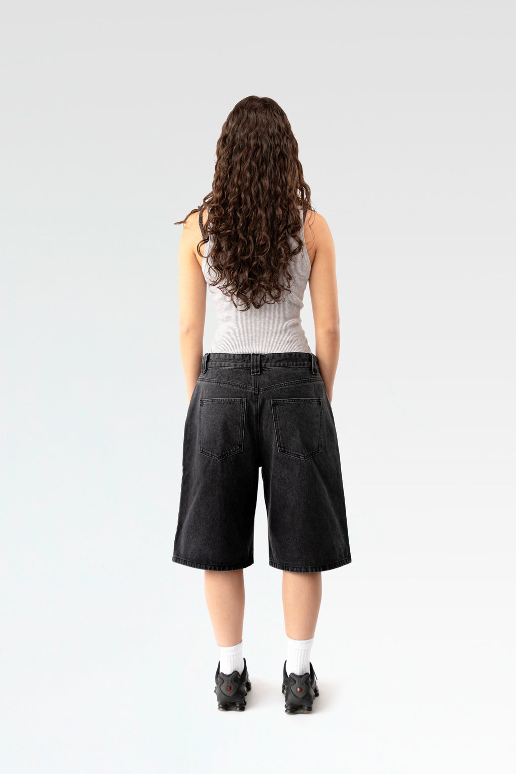 Rückansicht einer schwarzen, weit geschnittenen Jeansshorts aus gewaschenem Denim. Die Shorts reicht bis knapp übers Knie und zeigt zwei aufgesetzte Gesäßtaschen. Getragen von einer stehenden Person mit langen Locken, sportlichen Sneakers und weißem Tanktop.