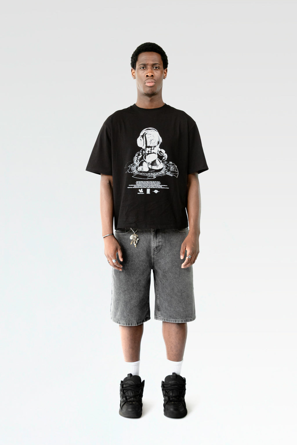 Frontale Studioaufnahme eines schwarzen T-Shirts mit weißem Roboterprint, getragen von einer männlich gelesenen Person. Der lockere Schnitt und das kontrastreiche Design sind klar erkennbar.