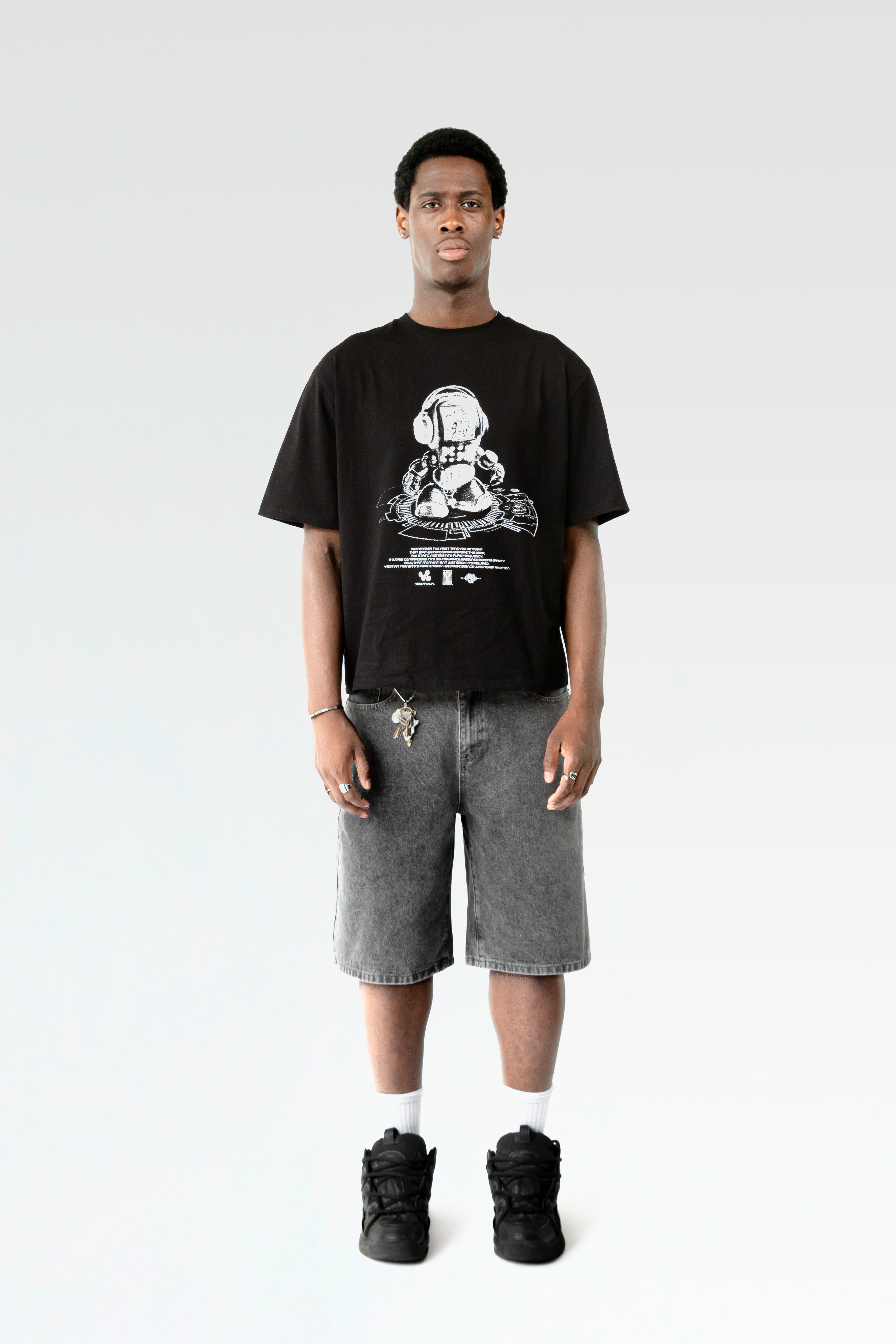Frontale Studioaufnahme eines schwarzen T-Shirts mit weißem Roboterprint, getragen von einer männlich gelesenen Person. Der lockere Schnitt und das kontrastreiche Design sind klar erkennbar.