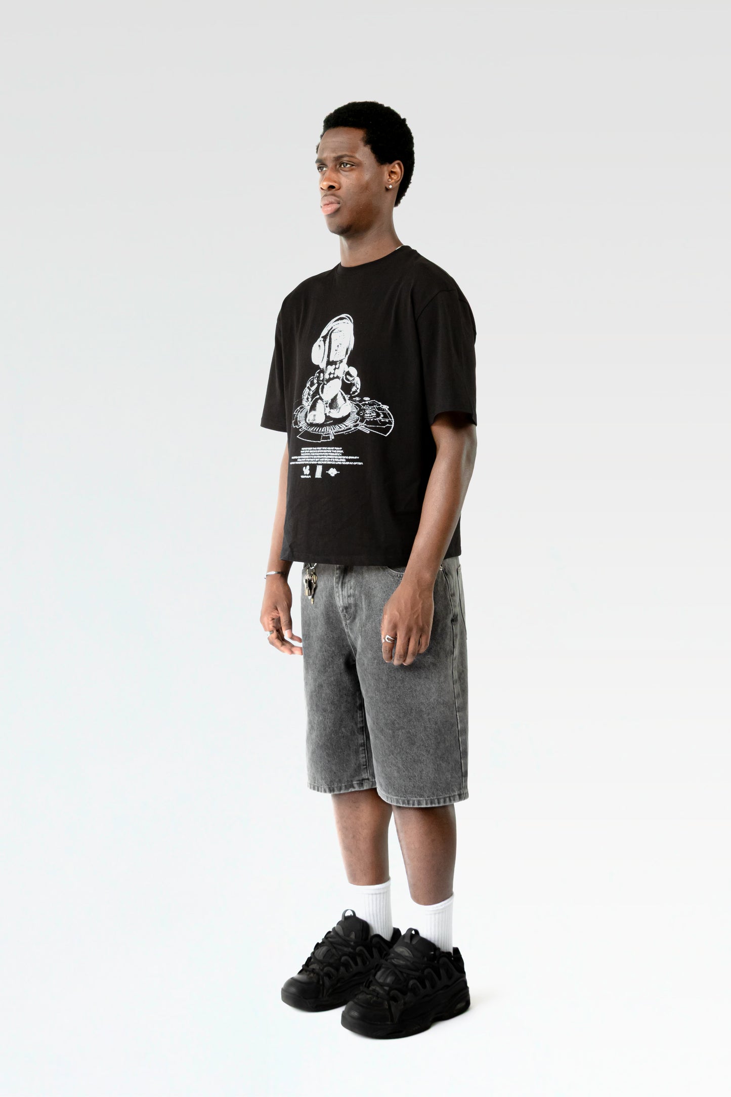 Seitliche Studioansicht des schwarzen T-Shirts mit weißem Motiv. Das Shirt fällt locker und wird mit einer hellgrau verwaschenen Bermuda-Shorts kombiniert.