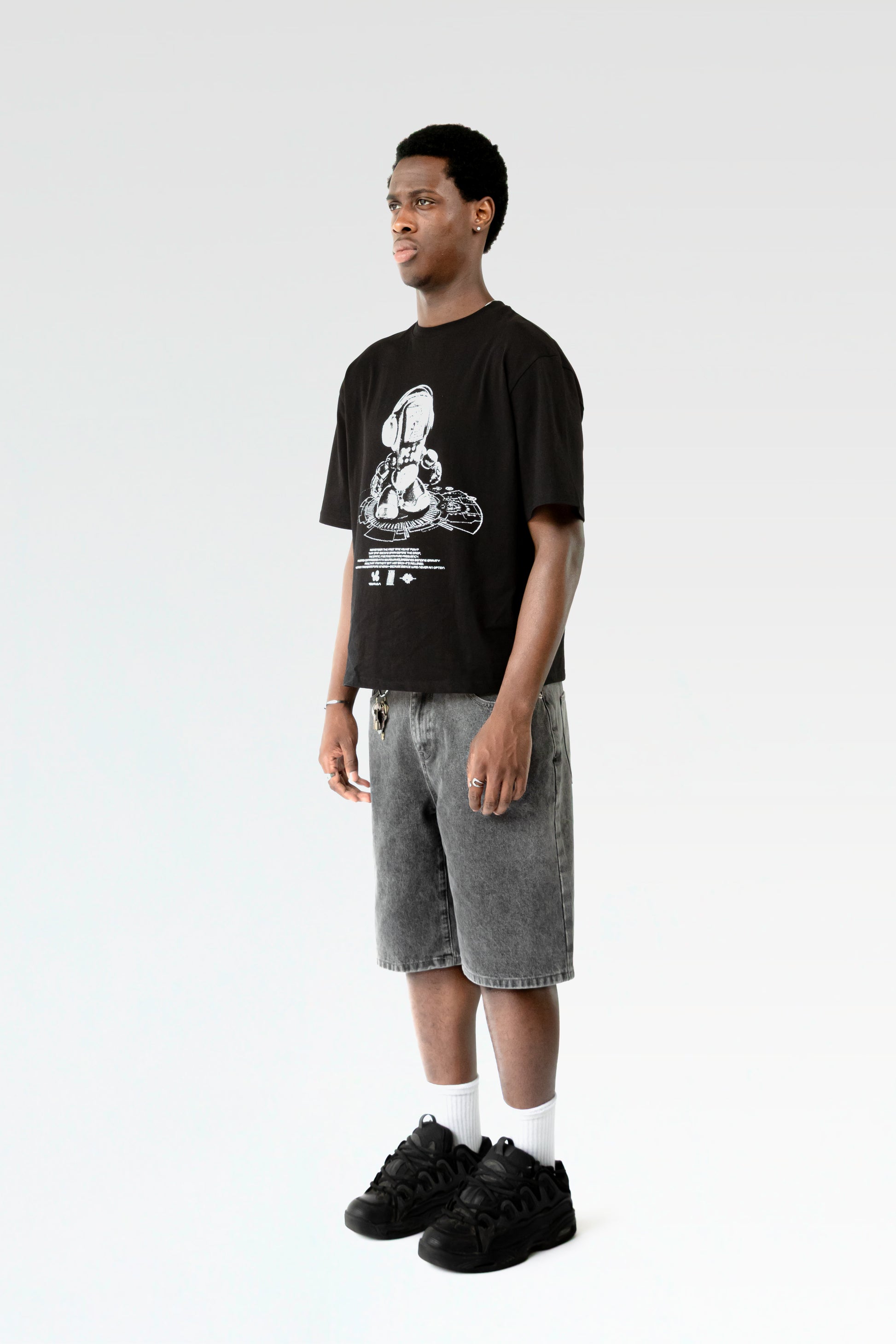 Seitliche Studioansicht des schwarzen T-Shirts mit weißem Motiv. Das Shirt fällt locker und wird mit einer hellgrau verwaschenen Bermuda-Shorts kombiniert.