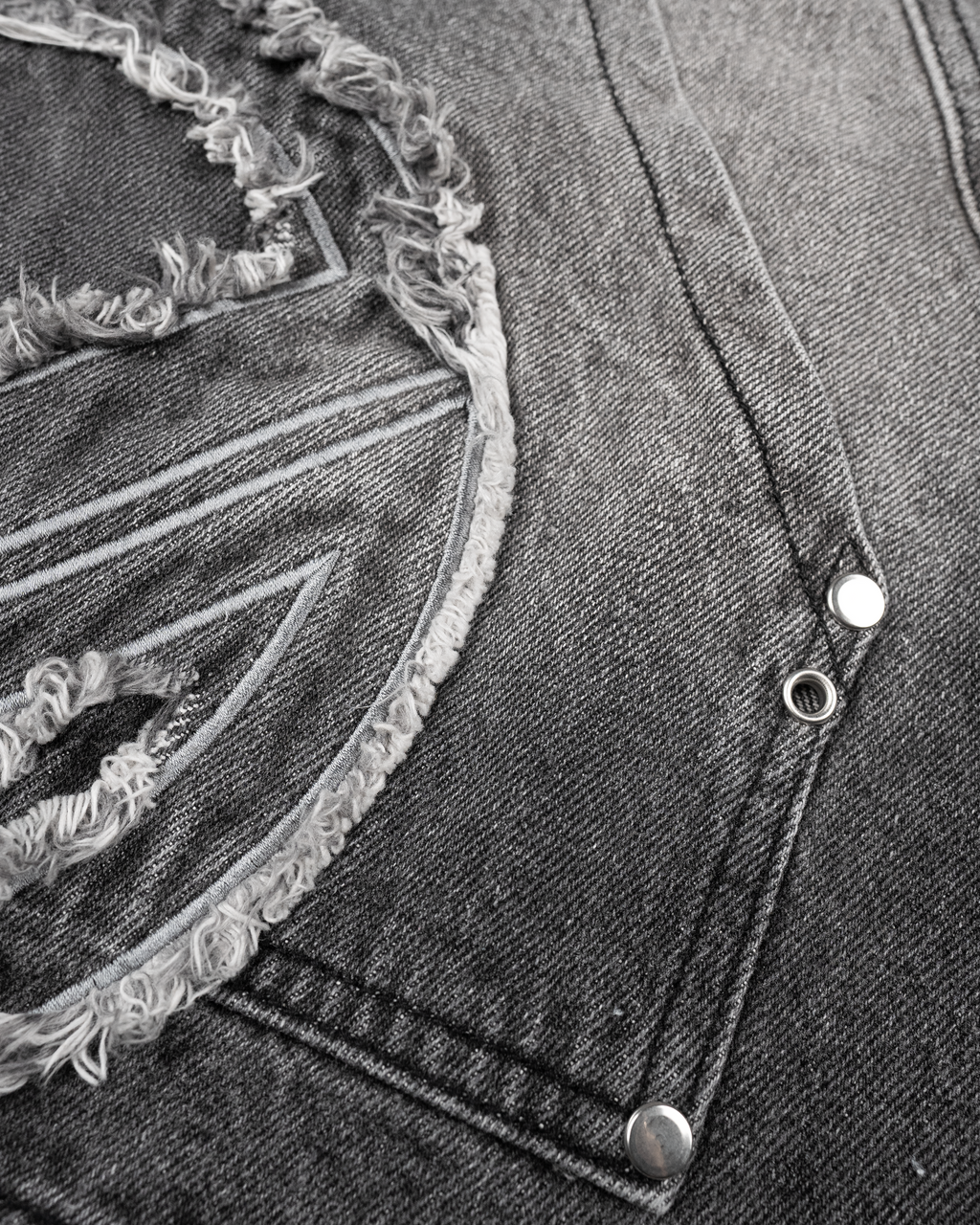 Nahaufnahme der linken Oberschenkeltasche der grauen Jeansshorts. Sichtbar sind der silberne Reißverschluss, silberne Nieten sowie das große Patch mit ausgefranster Kontur und kontrastierender Stickerei.