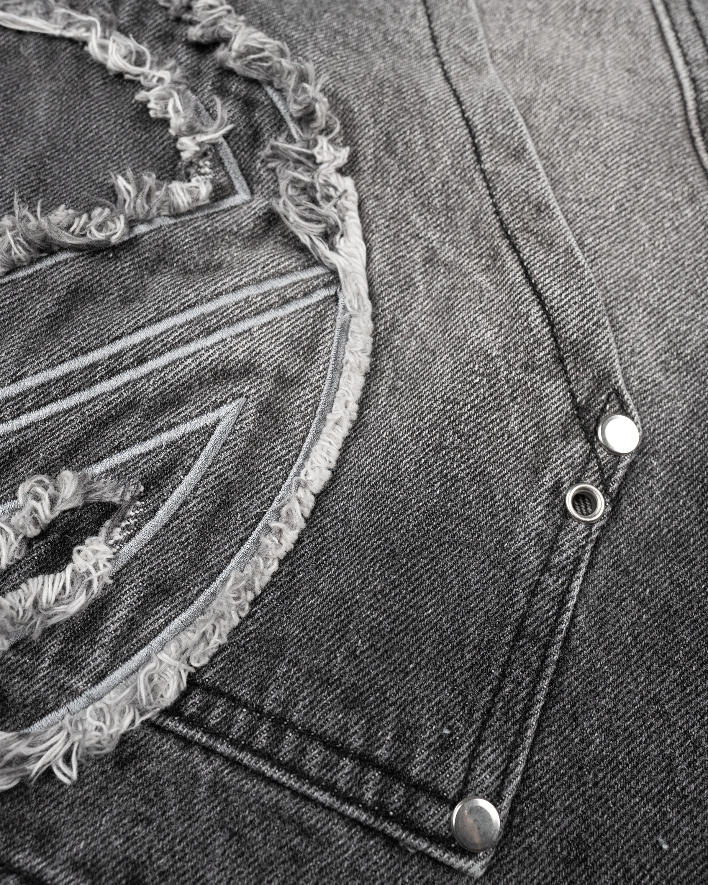 Nahaufnahme der linken Oberschenkeltasche der grauen Jeansshorts. Sichtbar sind der silberne Reißverschluss, silberne Nieten sowie das große Patch mit ausgefranster Kontur und kontrastierender Stickerei.