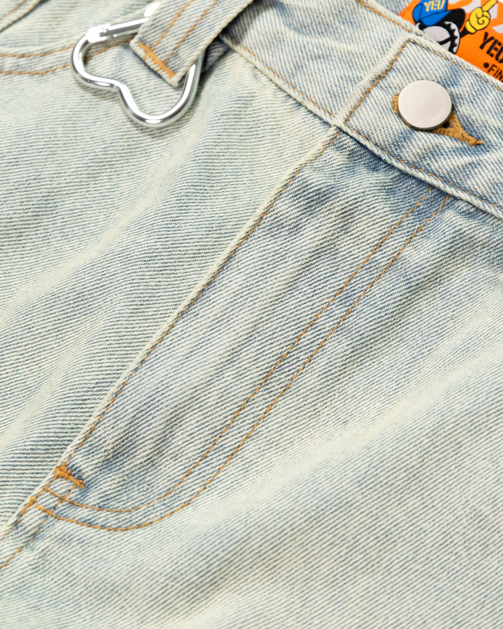 Detailaufnahme der Gürtelschlaufe mit silbernem Herz-Karabiner auf hellblauem Denim. Die grobe Stoffstruktur und Nahtverarbeitung sind sichtbar.