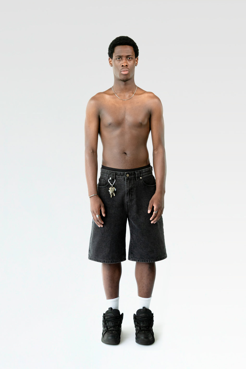 Frontale Studioaufnahme einer schwarzen Bermuda-Shorts an einer männlich gelesenen Person. Die Shorts endet knapp über dem Knie und ist mit einem Karabiner samt Schlüsselbund an der Gürtelschlaufe ausgestattet.