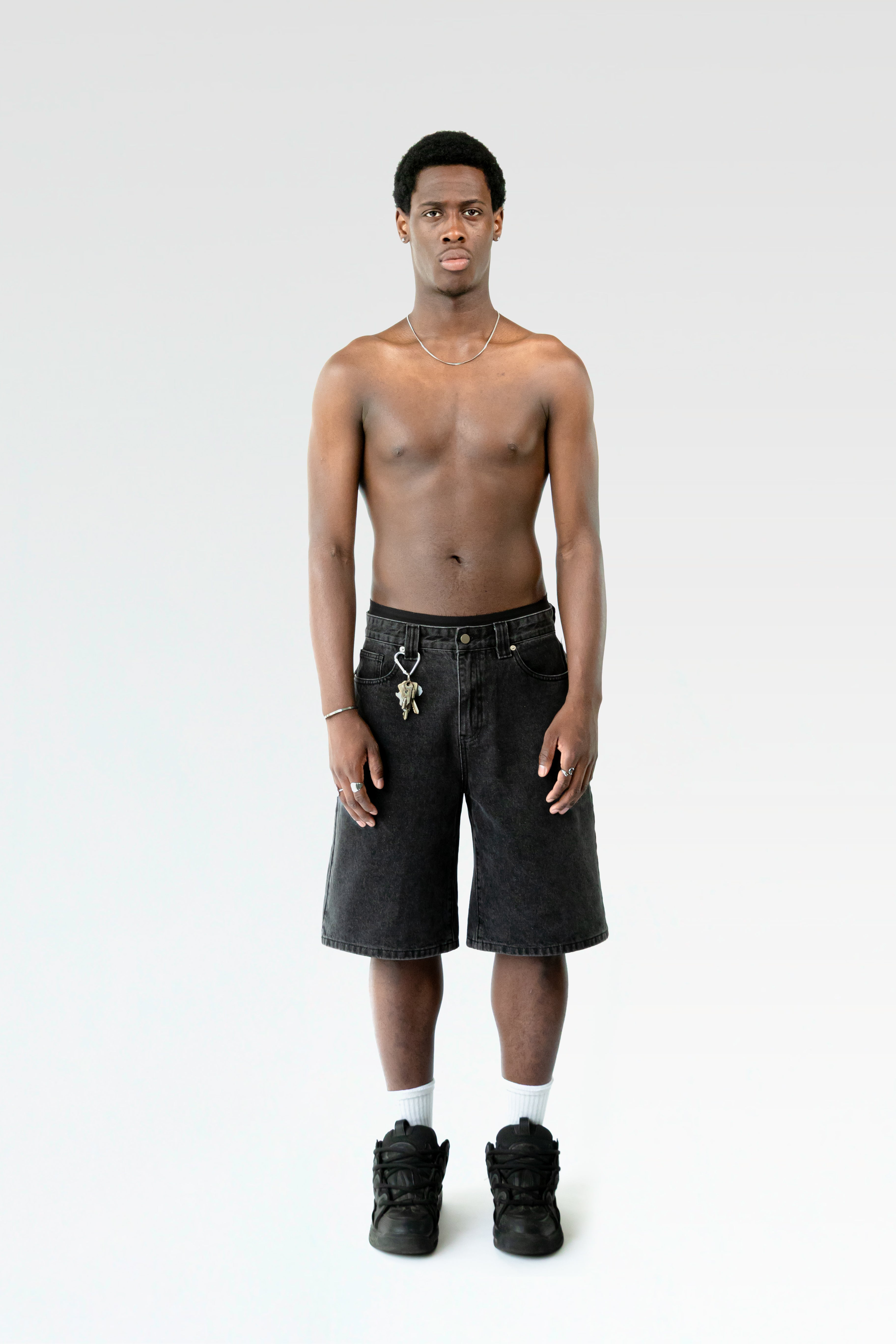 Frontale Studioaufnahme einer schwarzen Bermuda-Shorts an einer männlich gelesenen Person. Die Shorts endet knapp über dem Knie und ist mit einem Karabiner samt Schlüsselbund an der Gürtelschlaufe ausgestattet.