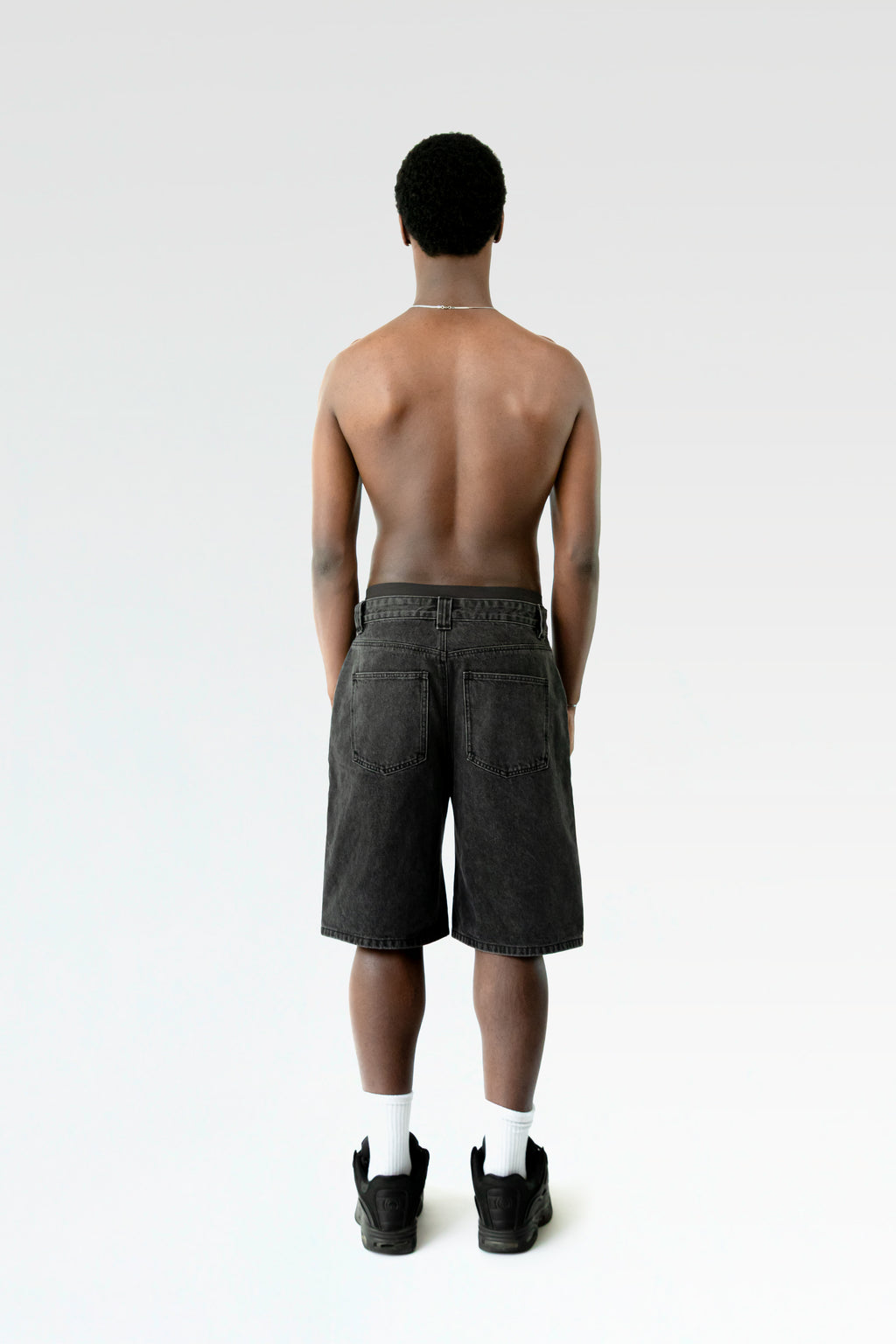 Rückansicht der schwarzen Bermuda-Shorts an einer stehenden Person. Die Hose hat zwei aufgesetzte Gesäßtaschen und einen geraden Saum.